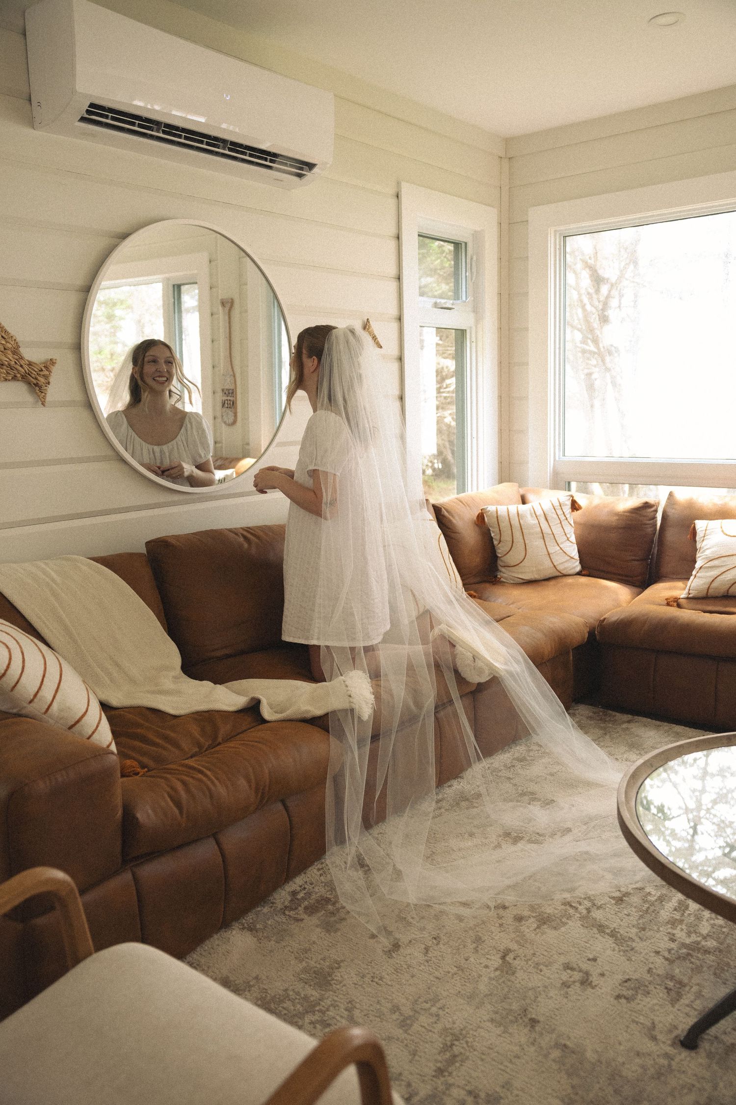 Bride in white wedding gown and long veil admires her reflection in a round mirror in a cozy living room.