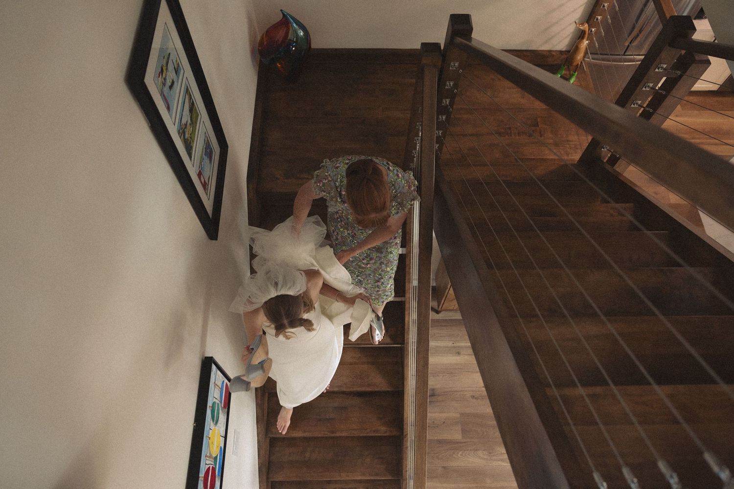 Overhead view of bride in white gown descending wooden staircase with family member.