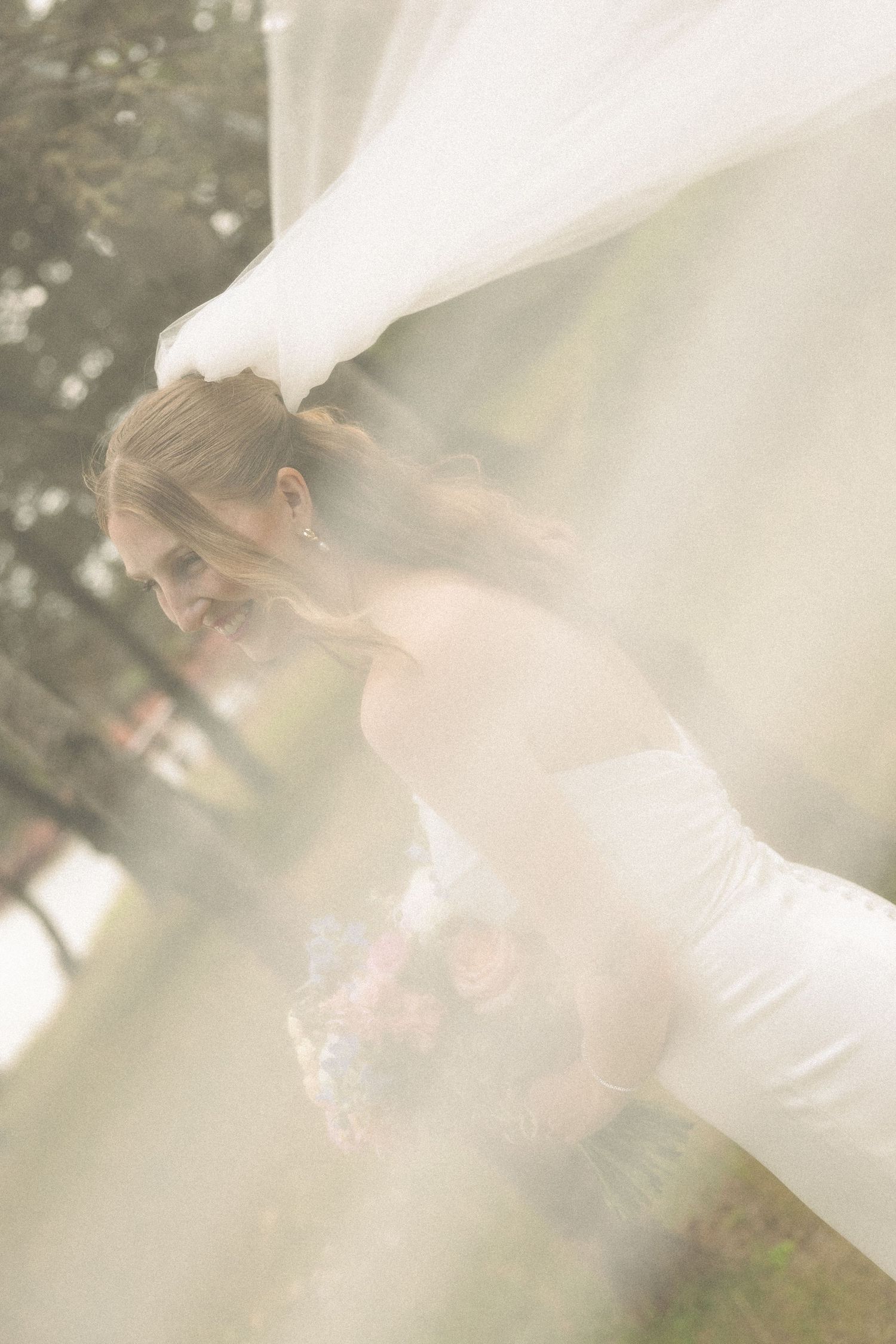 Bride in white gown smiling as her veil billows around her outdoors.