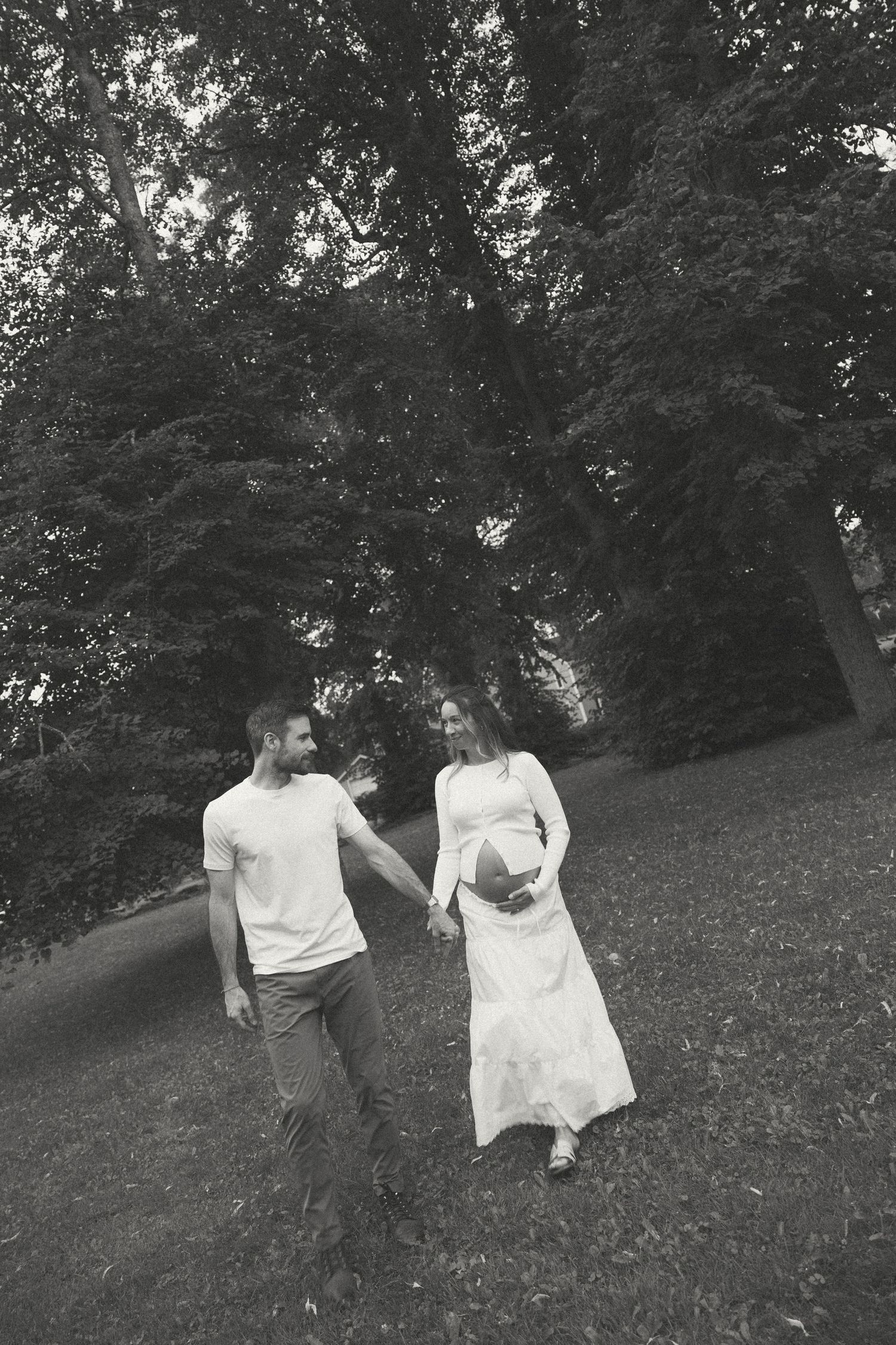 Black and white photo of pregnant couple holding hands walking under tall trees.