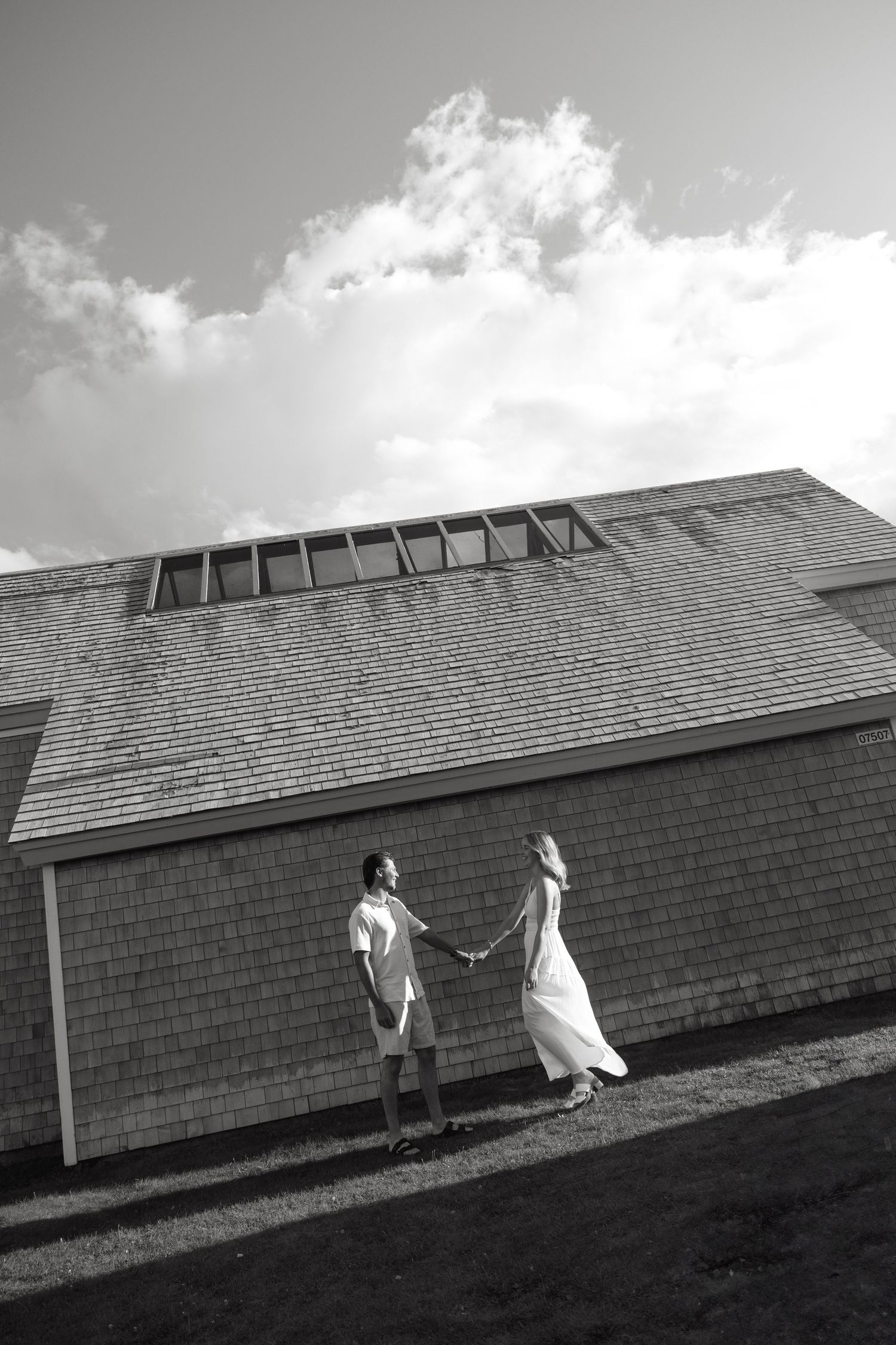 Black and white photo of a couple holding hands in front of a shingled building under a cloudy sky.