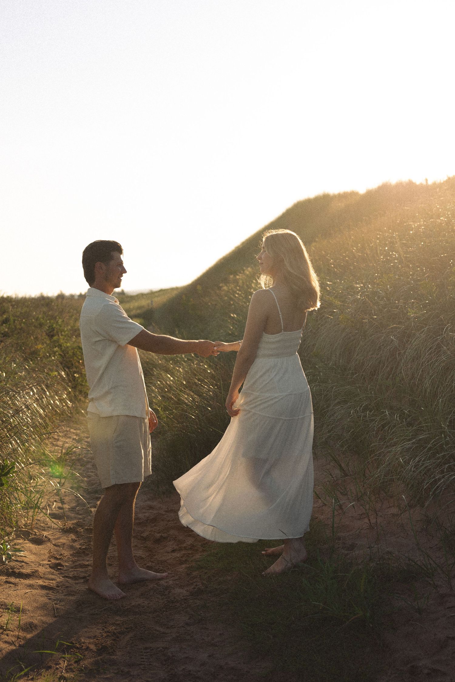 Couple holding hands at sunset on sandy dune path with tall grass.