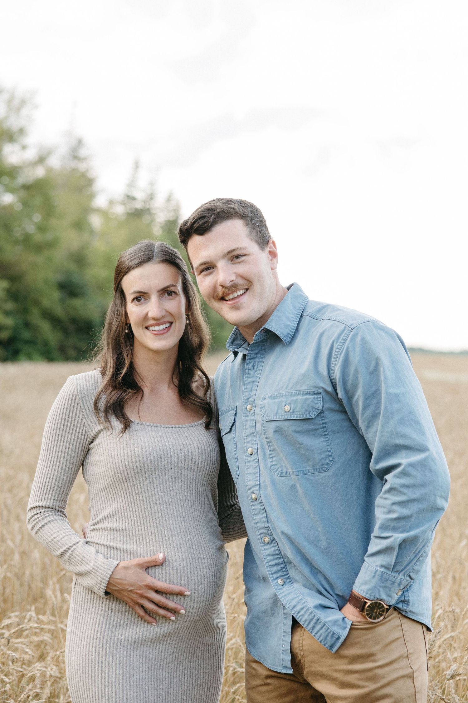 Smiling pregnant couple posing together in a golden wheat field, woman in gray dress cradling baby bump.