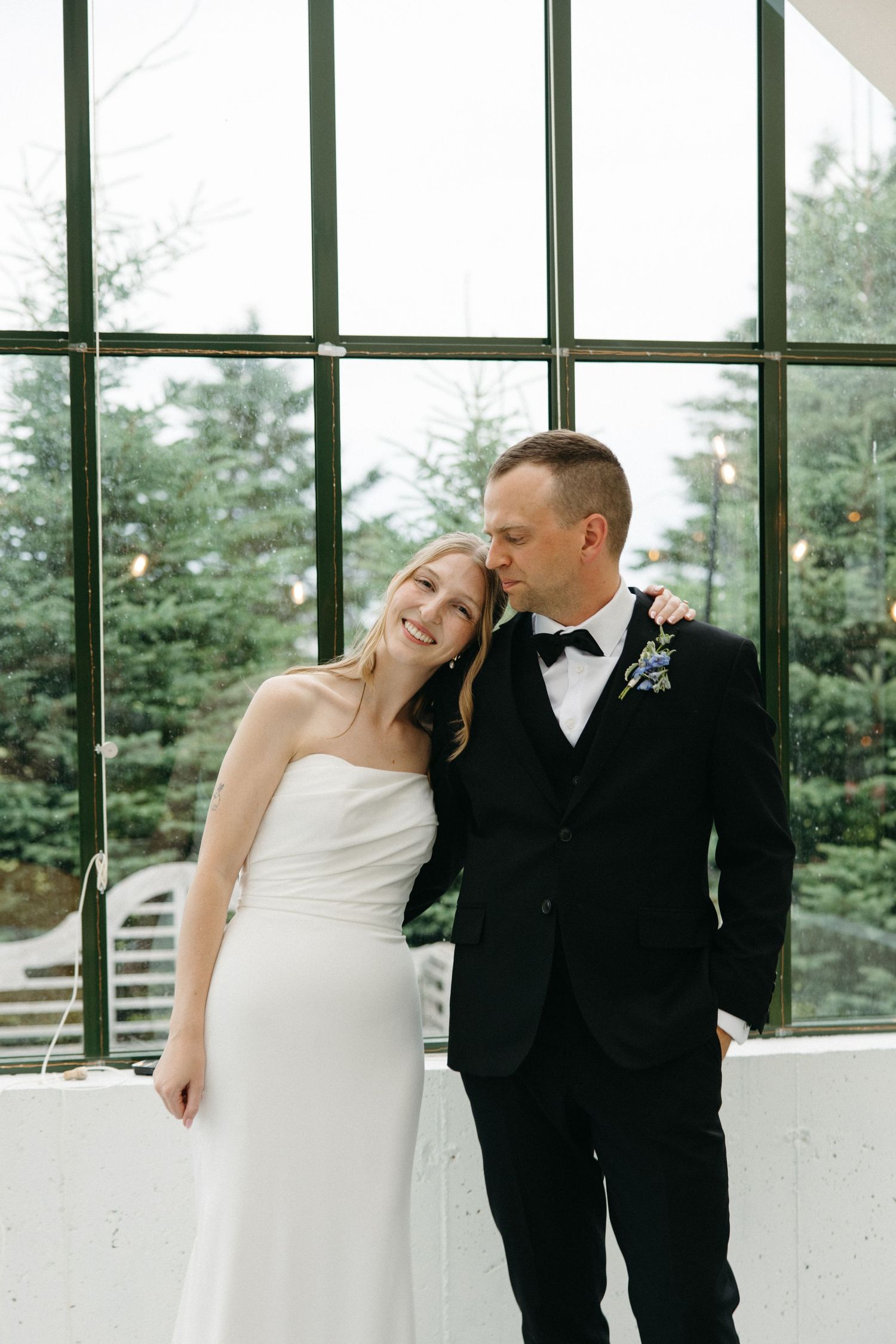 Bride leaning on groom in black tuxedo smiling before large window with evergreen trees.