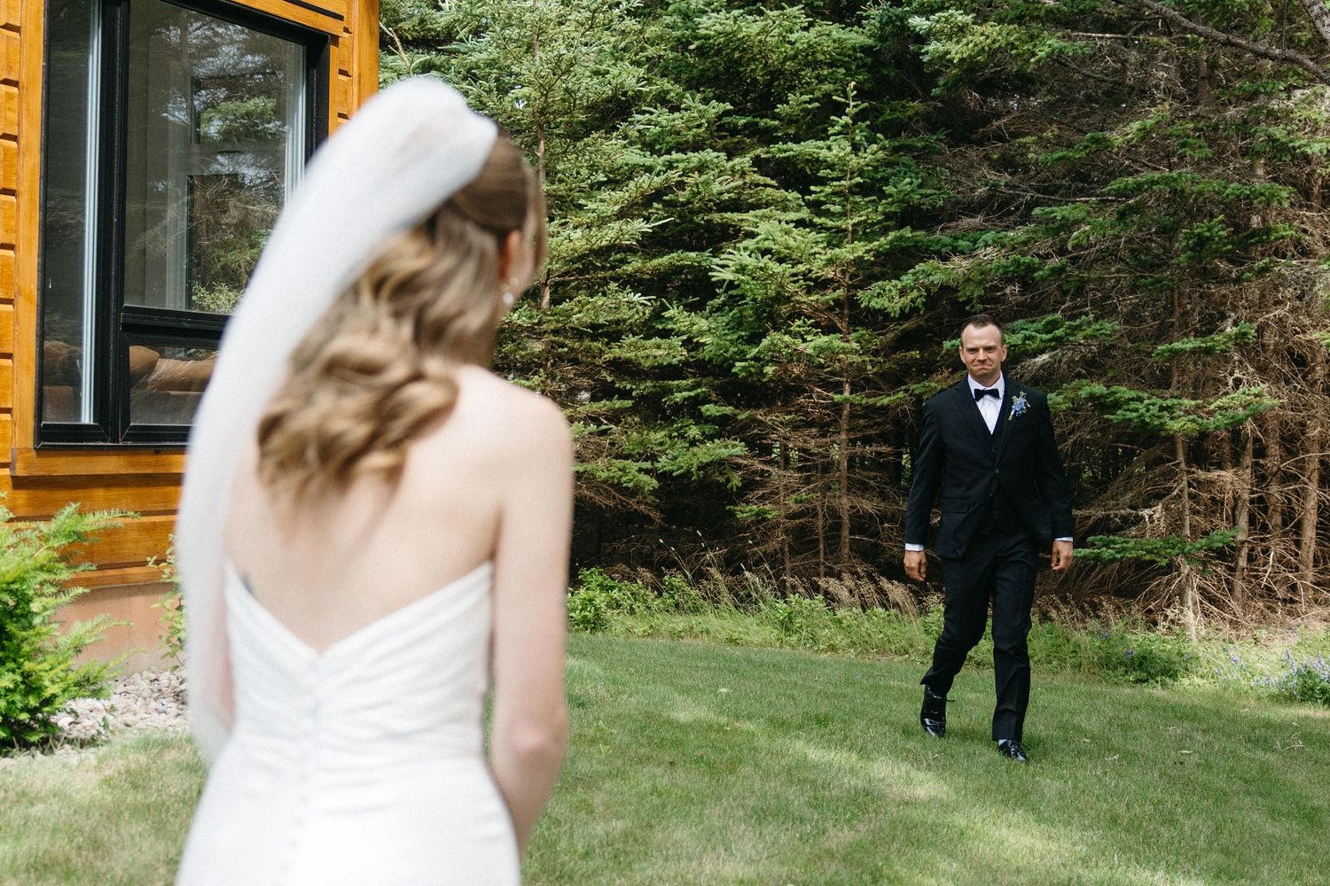 Bride in white strapless gown and veil stands outdoors as groom in black tuxedo approaches on green lawn.