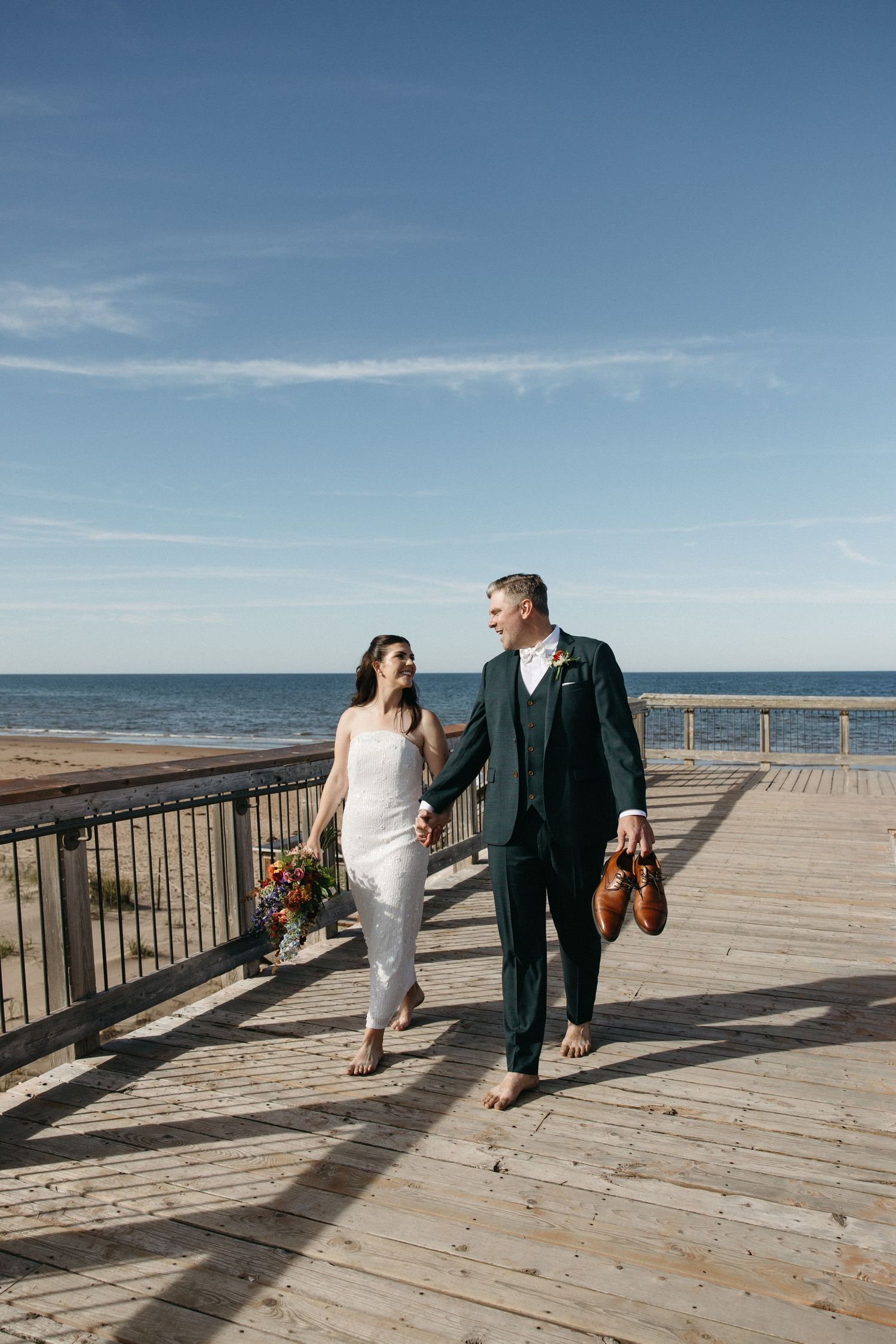 Barefoot bride in a white dress and groom in a green suit holding hands and shoes while walking along a sunny seaside boardwalk.