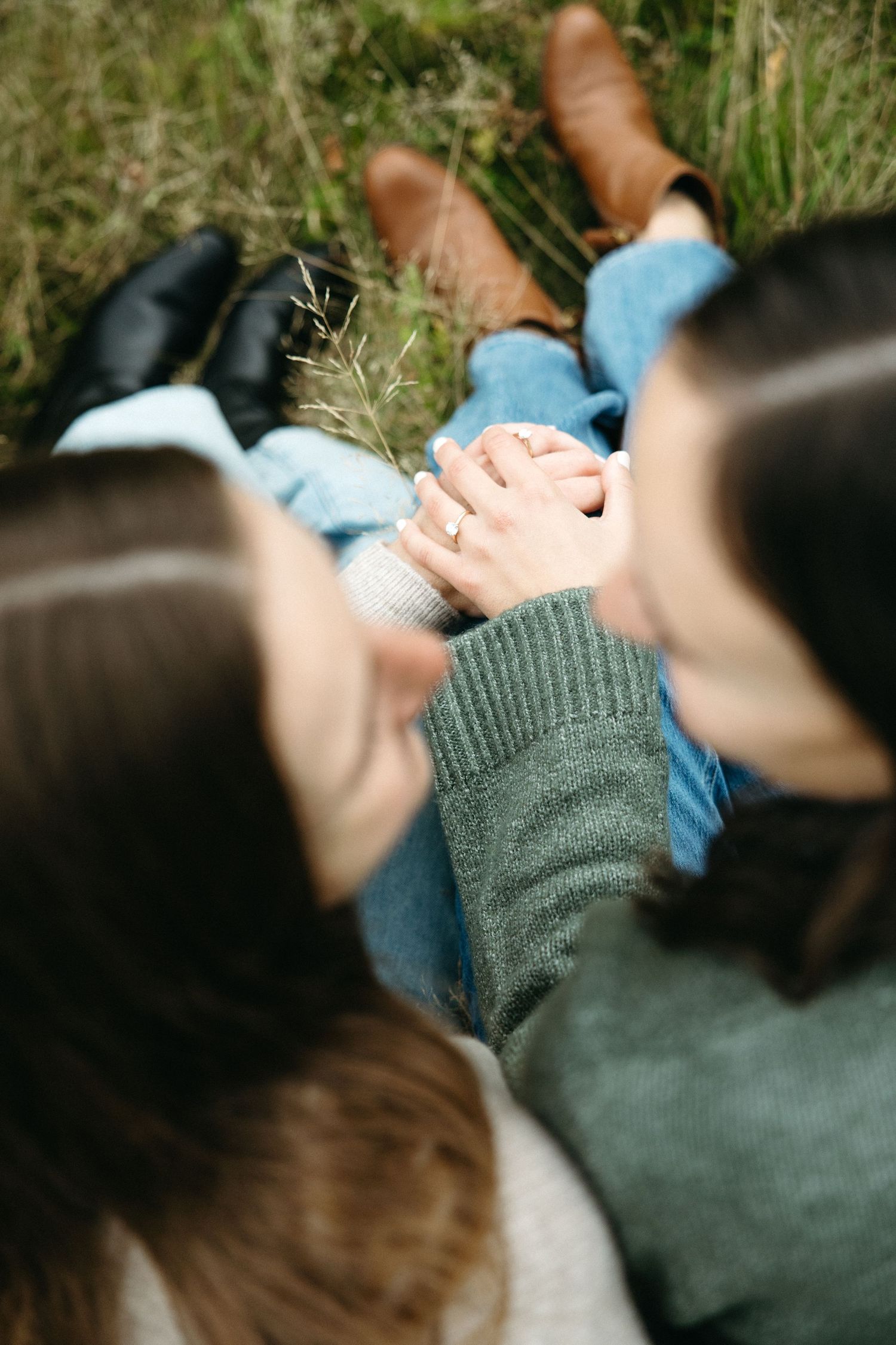 Overhead view of two women sitting in the grass holding hands, wearing jeans and boots, with rings visible on their fingers.