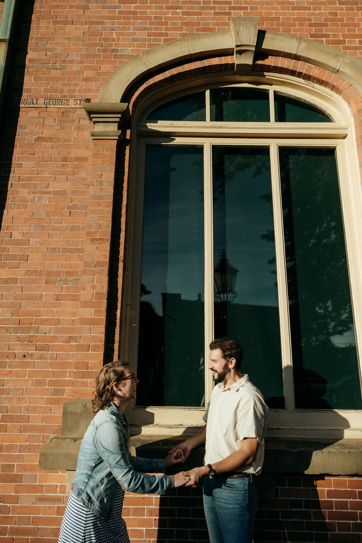 A couple interacts playfully in front of an arched doorway of a brick building.