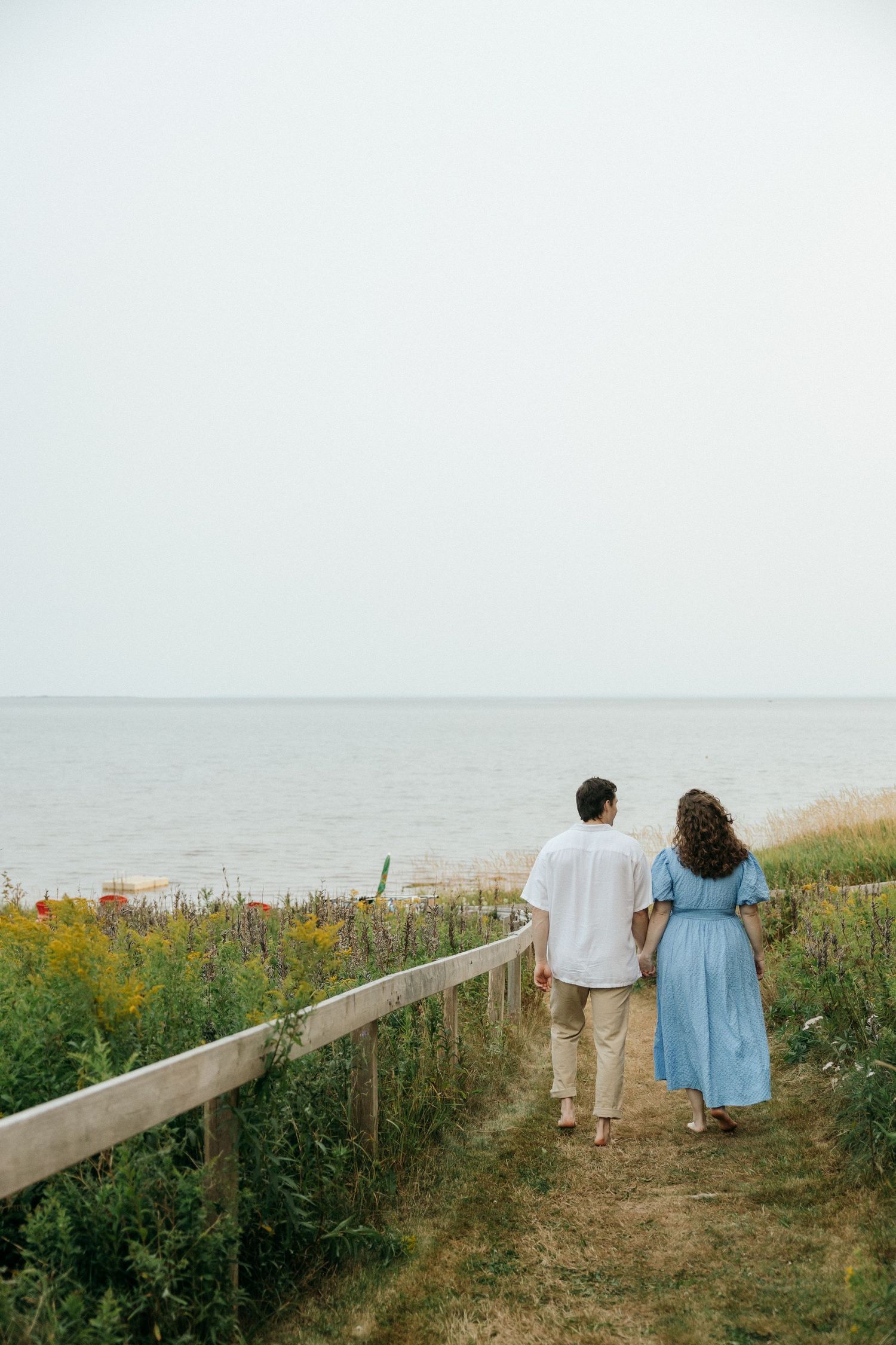 A couple strolling down a coastal path lined with wildflowers and wooden railings at sunset.