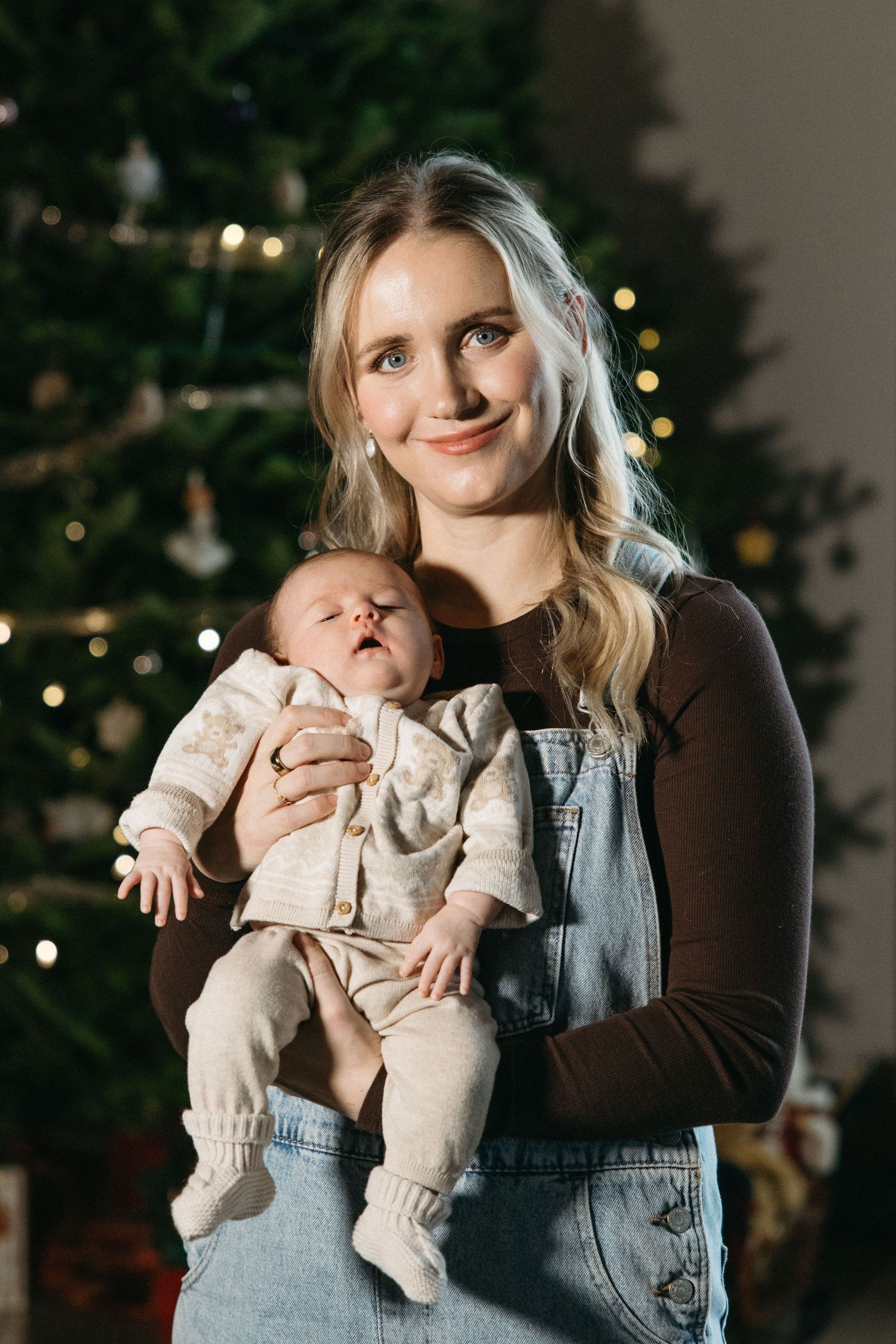 A baby in cozy winter clothes is held against backdrop of twinkling Christmas tree lights.