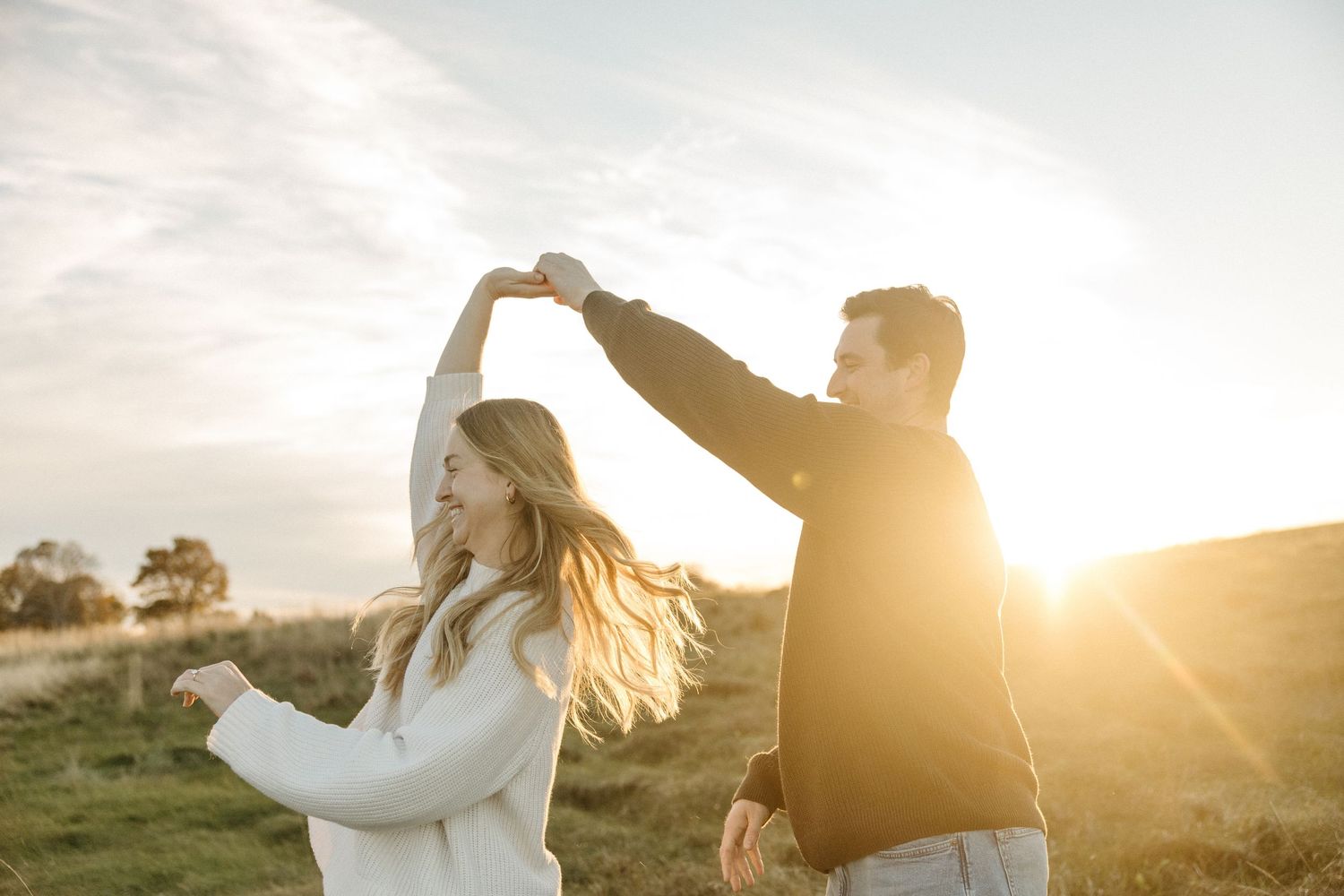 A smiling couple holds hands and dances on a grassy hillside at golden hour as warm sunset light glows behind them.