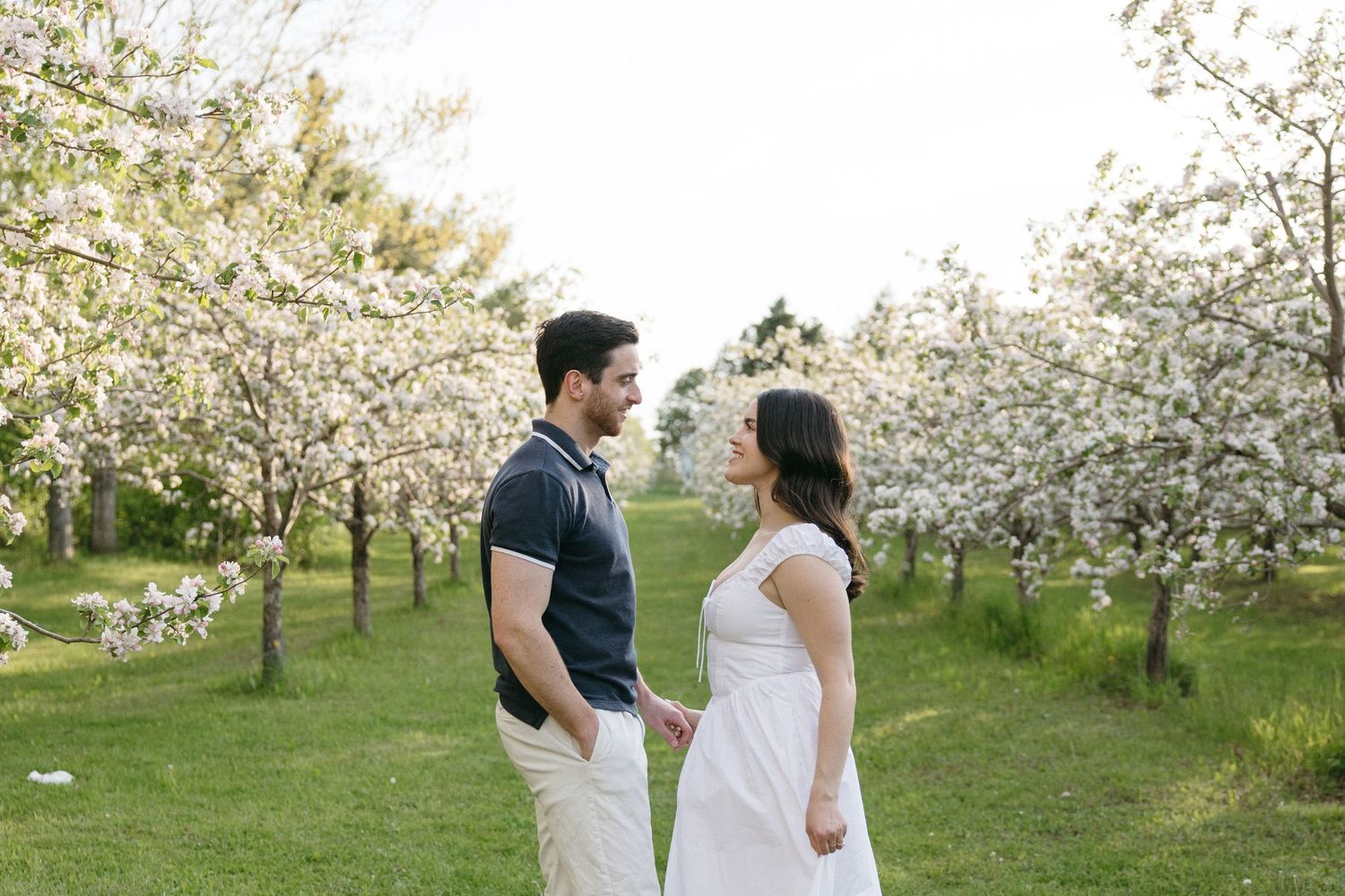 Couple holding hands and facing each other among white flowering trees in a lush green orchard.