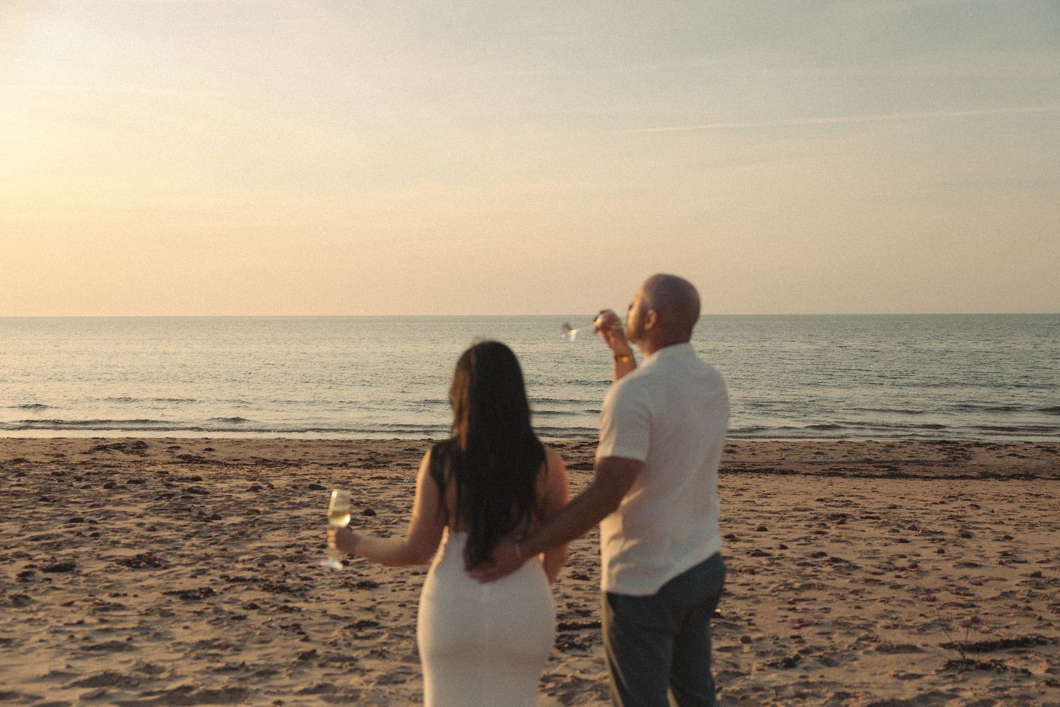A couple toasts with champagne glasses on a beach while watching a romantic sunset together.