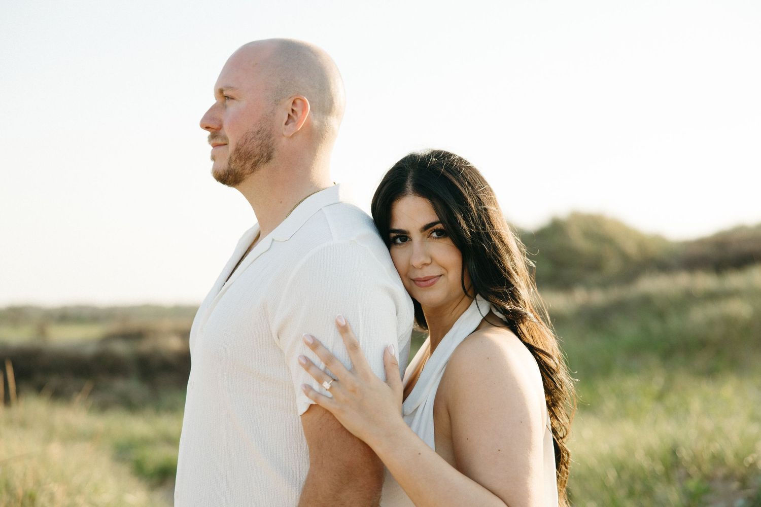 Couple posing outdoors in a sunny field, woman in white dress embracing man from behind, looking at camera.
