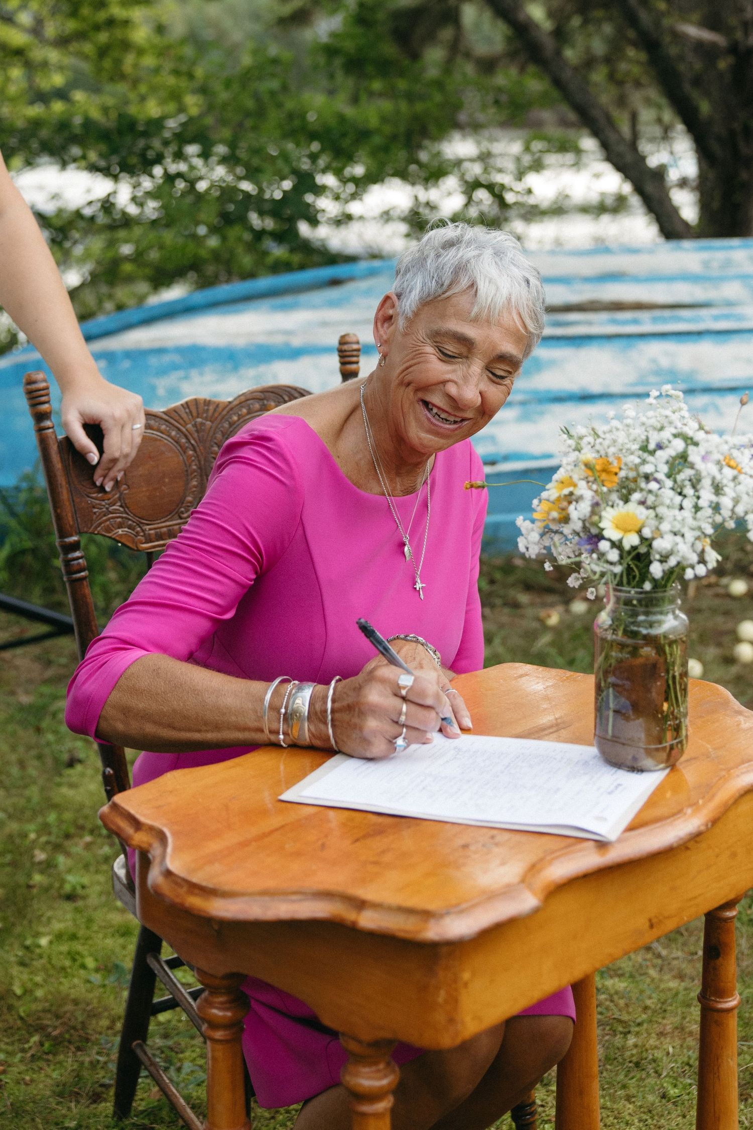 Person in pink shirt writing at a rustic wooden table outdoors with flowers in a mason jar.