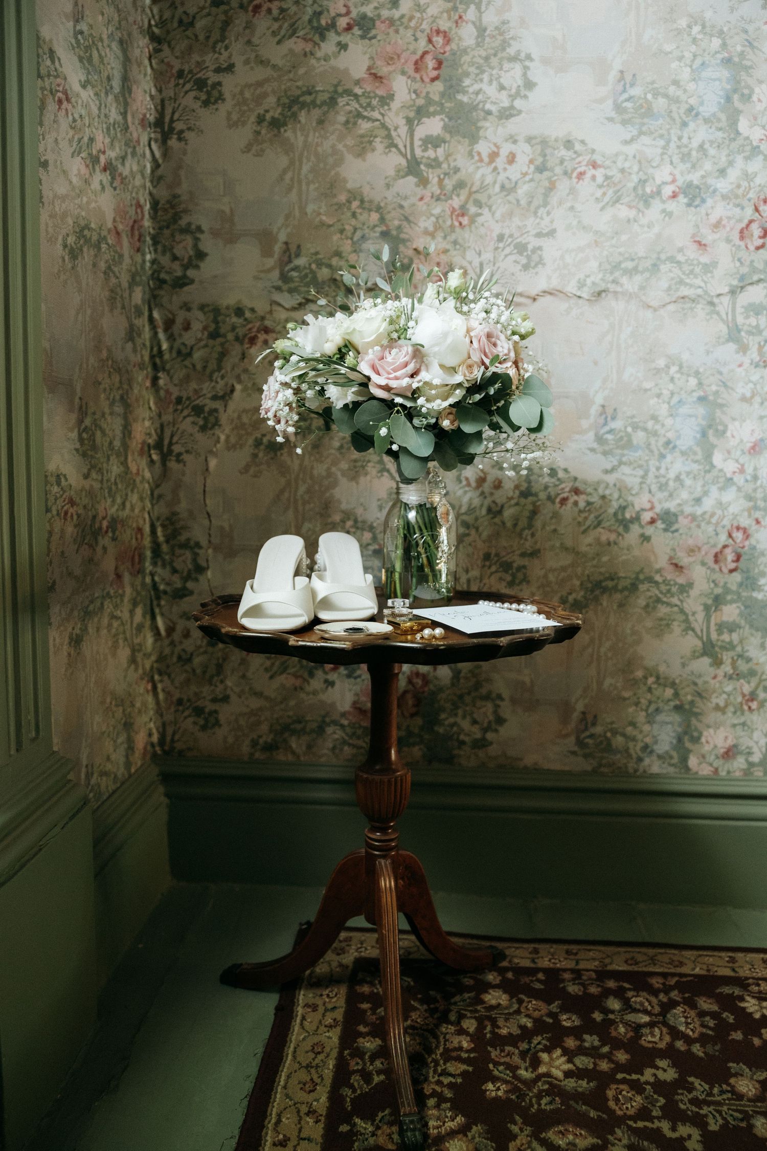 Vintage-inspired wedding details on a round table against floral wallpaper in moody lighting.