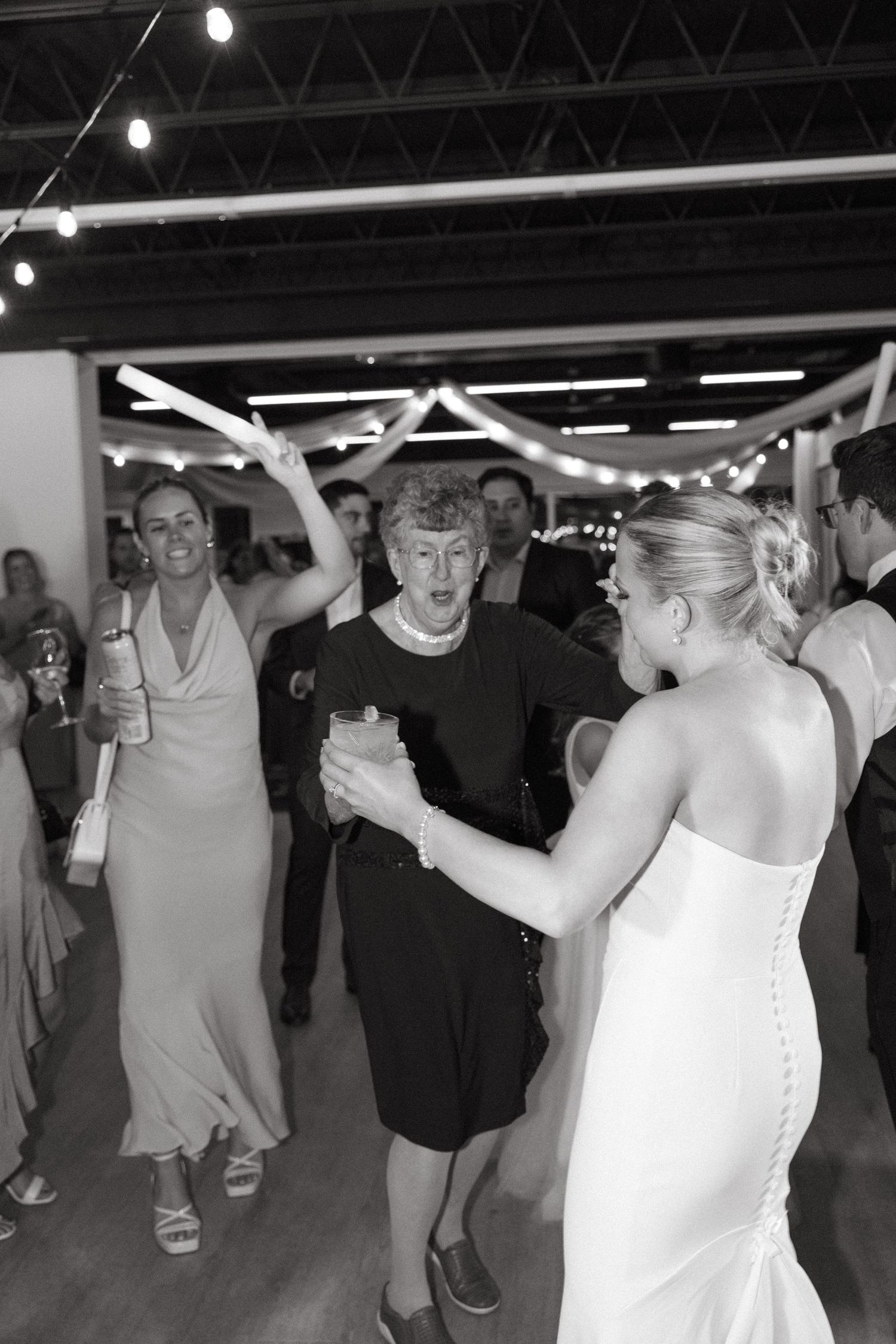 People dance energetically at a wedding reception under industrial ceiling and string lights.