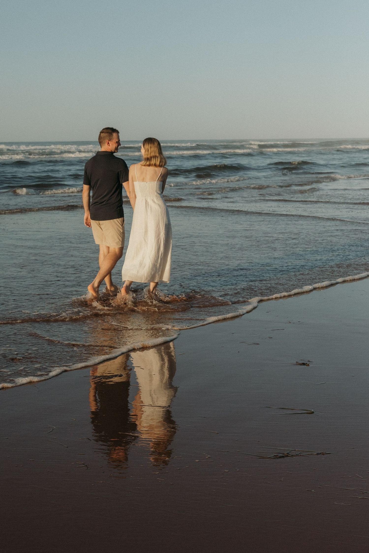 A couple walks through shallow ocean waves as their reflection appears in the wet sand at sunset.