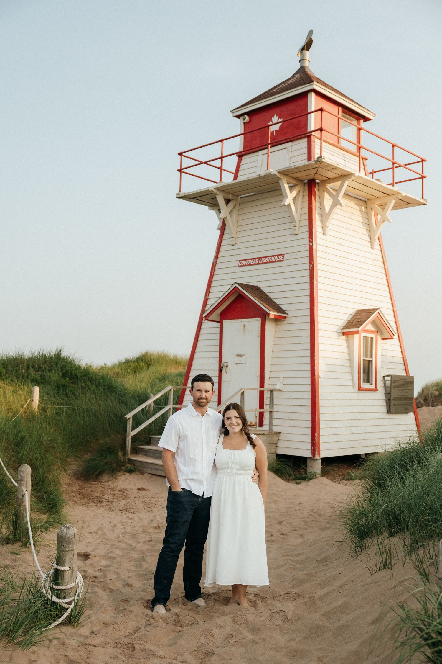 A couple poses in front of a white and red lighthouse on a sandy beach.