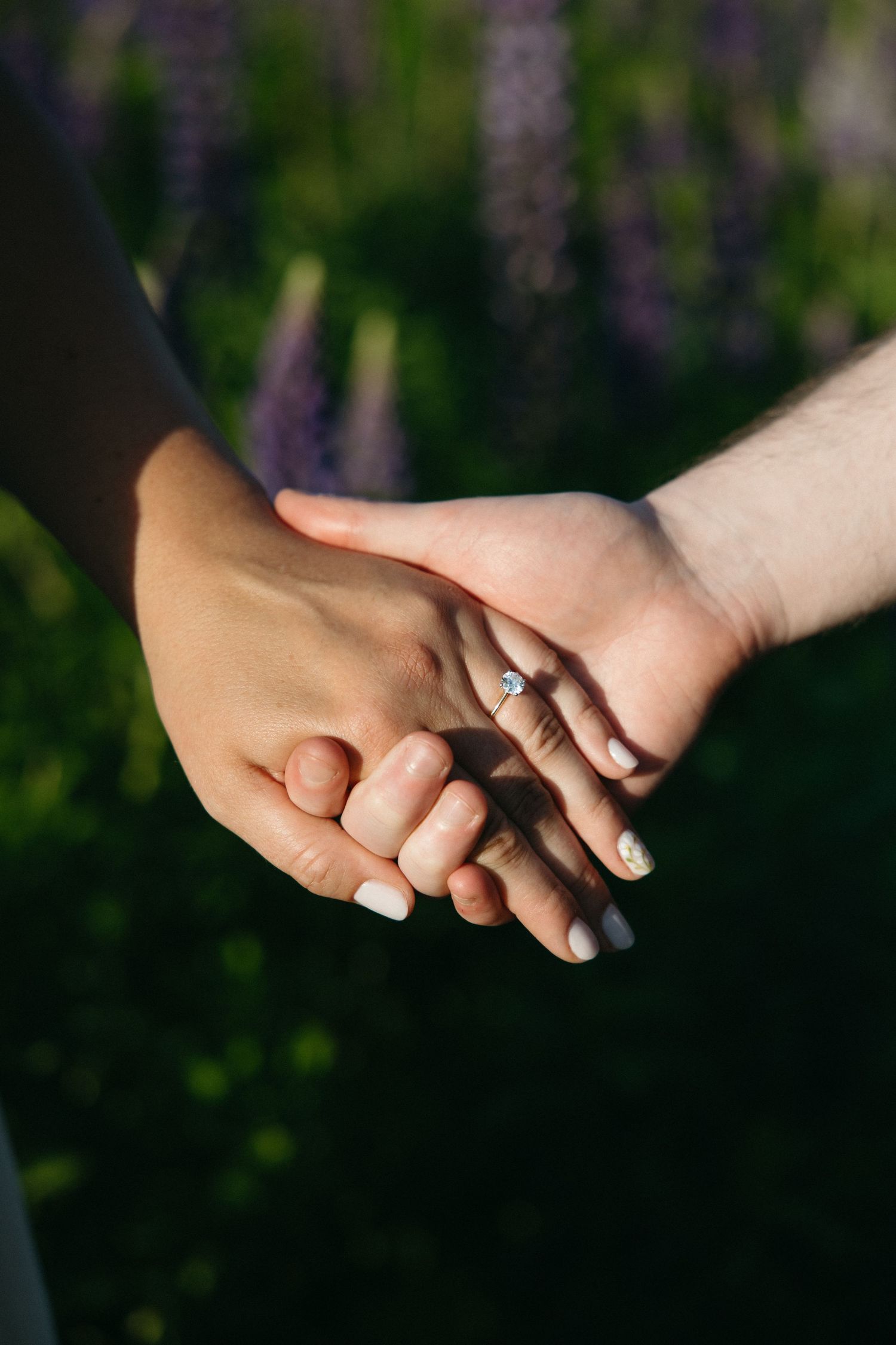 Close-up of couple holding hands with engagement ring in flower field.