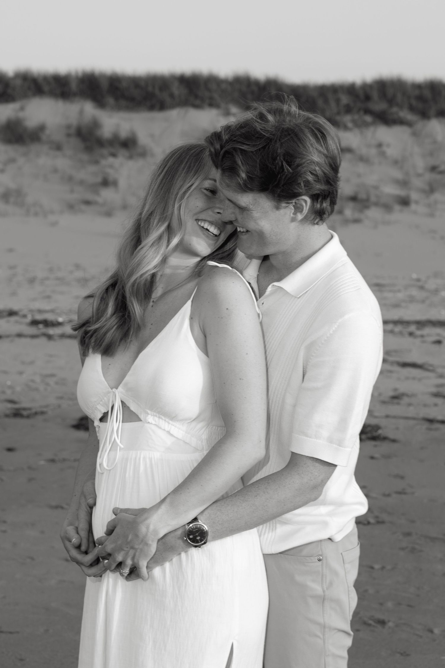 Black and white photo of romantic couple hugging on sandy beach, woman in white dress smiling joyfully.