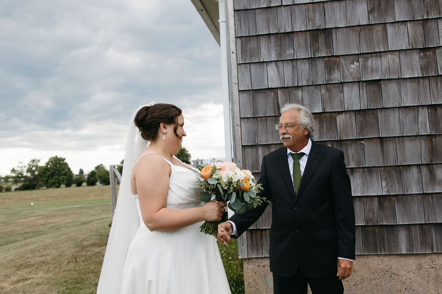 Bride in white gown holding a peach bouquet standing with her father before the wedding ceremony outdoors.