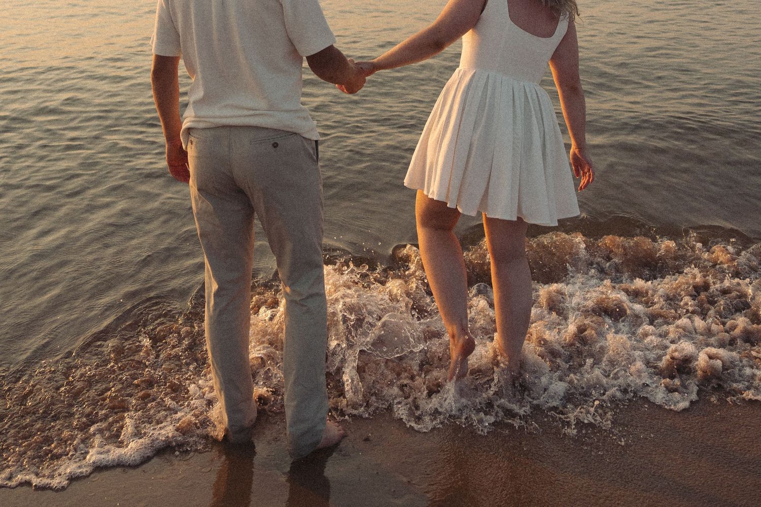 Couple holding hands walking barefoot on a sandy beach at sunset, waves washing over their feet.