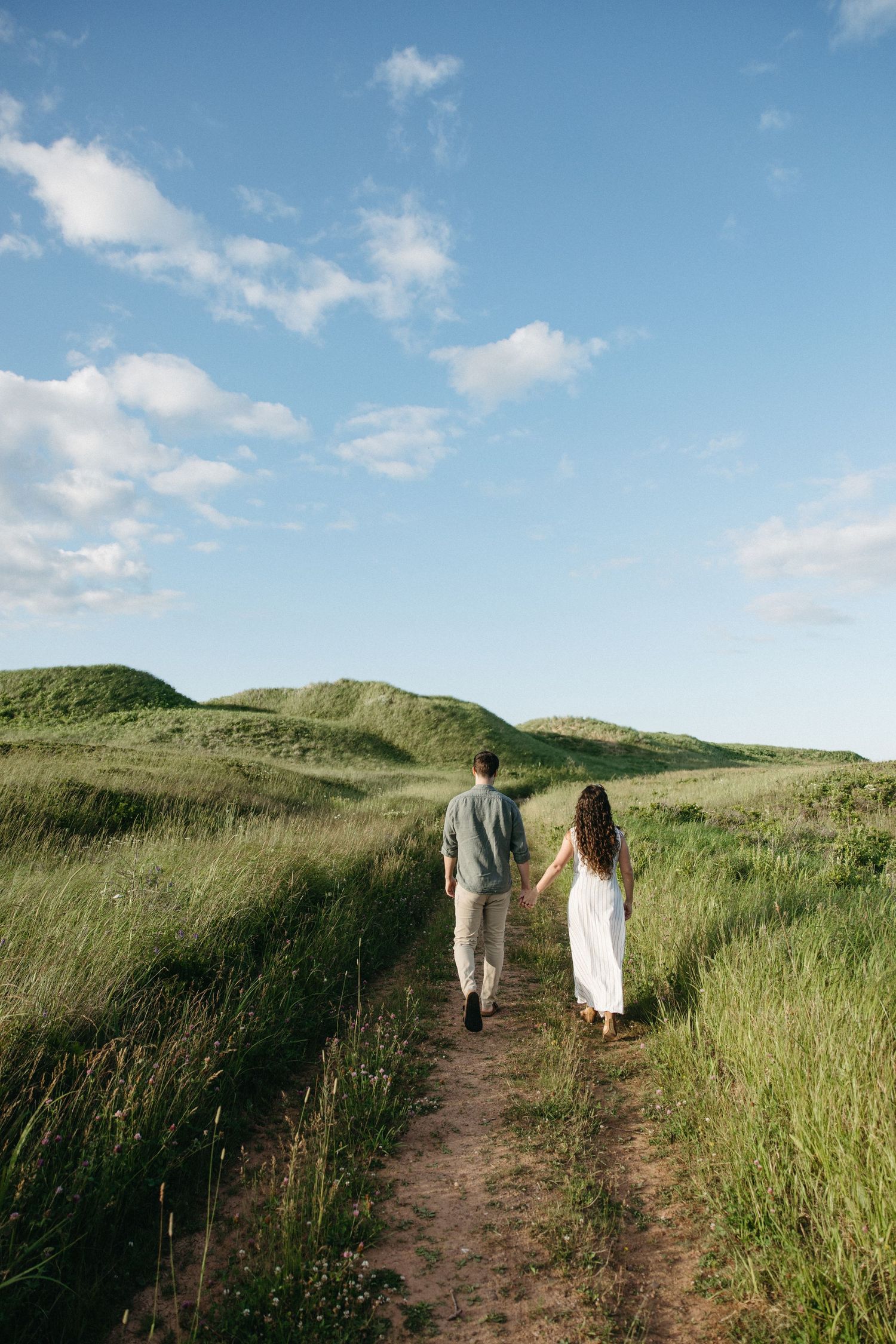 A couple walks hand-in-hand along a narrow dirt path through lush green grassy dunes under a blue sky.