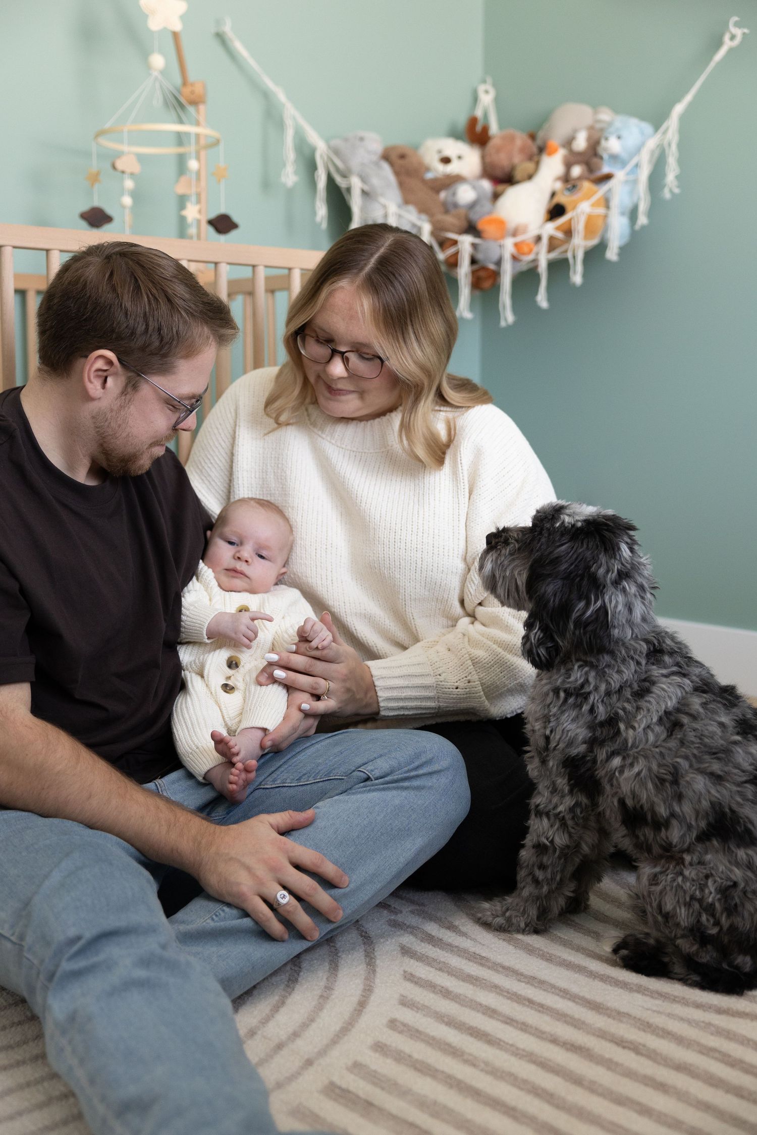 A family sits on the floor of a nursery with their dog while stuffed animals hang in the background.