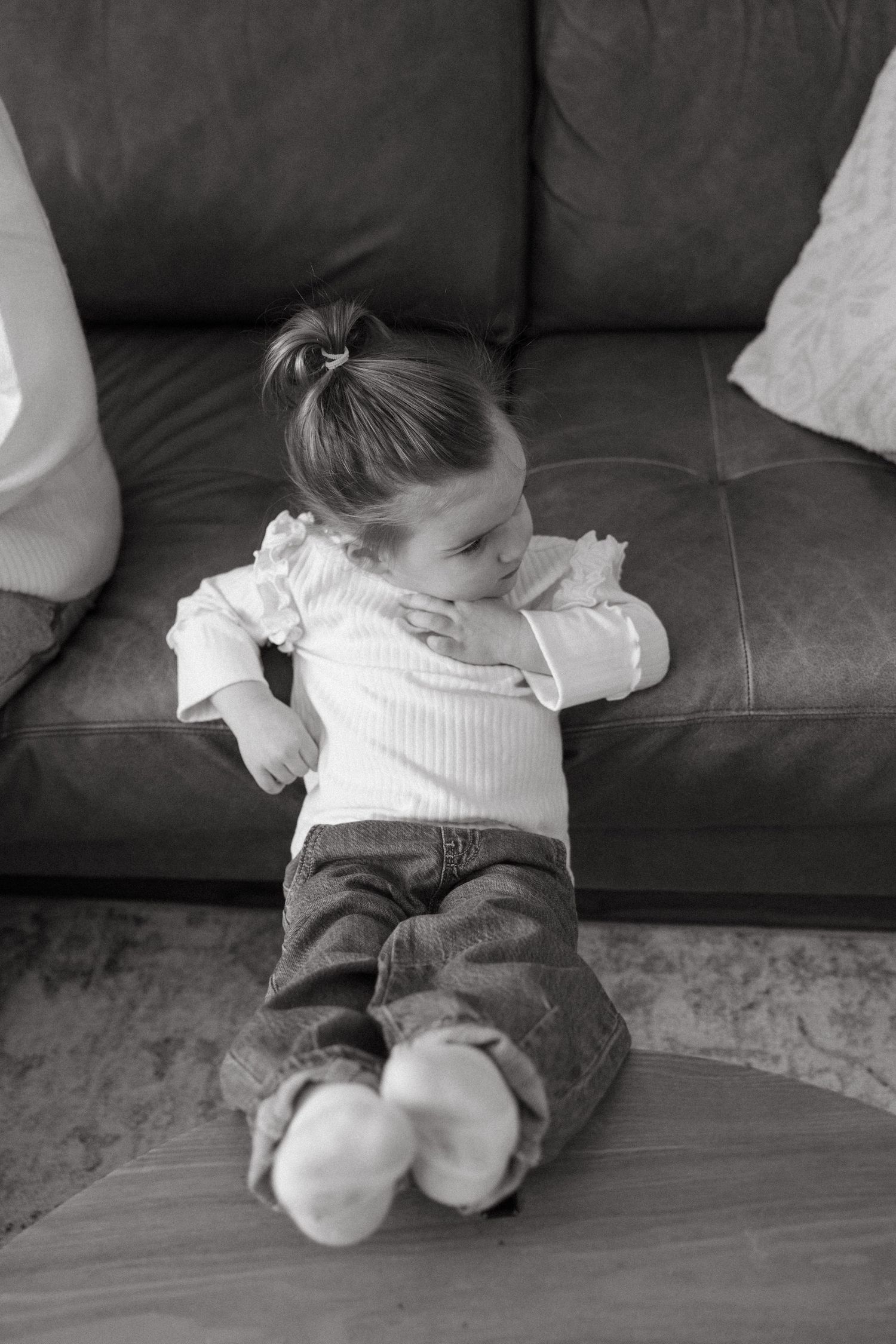 A small child in jeans and a white sweater lounges on a dark couch in black and white.