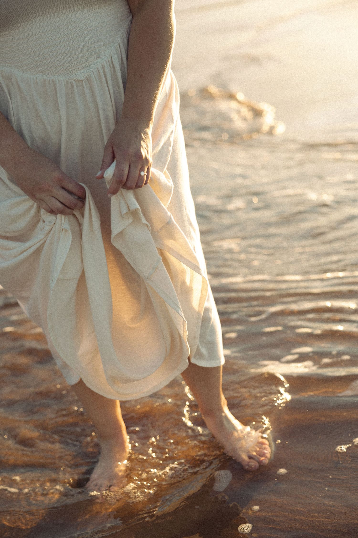 A woman in a white dress lifts her hem while wading barefoot in golden ocean waves at sunset.