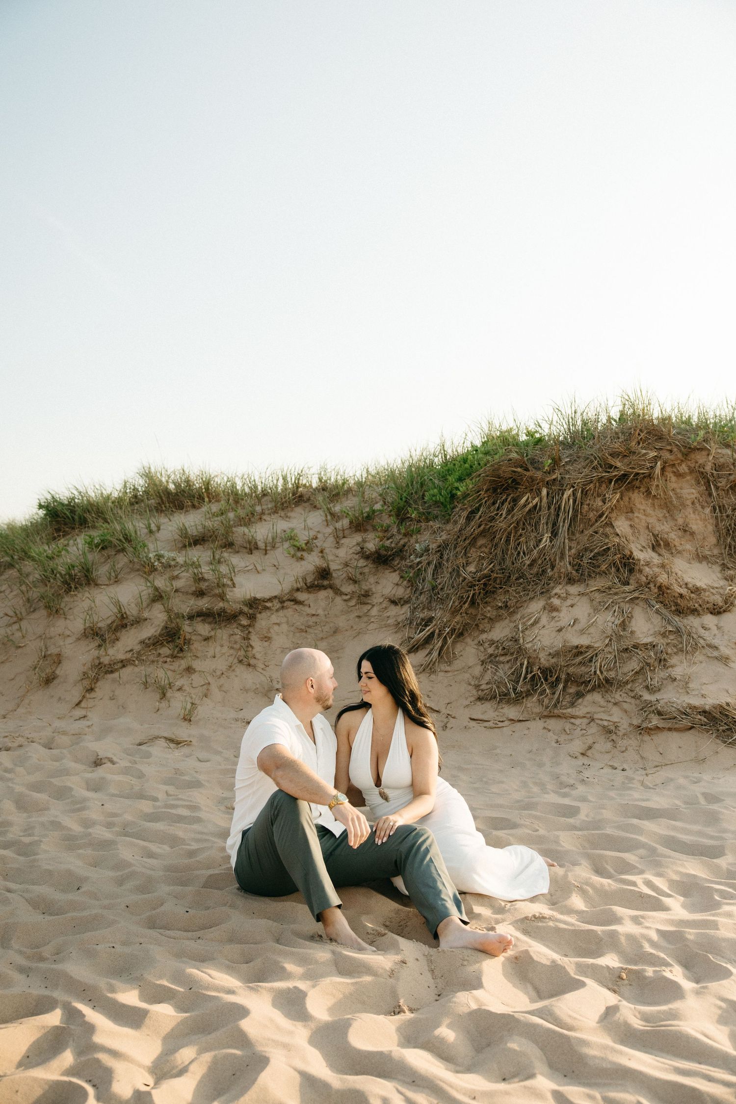 Engaged couple sitting together on sandy beach dunes, smiling at each other, woman in white dress, man in casual attire.