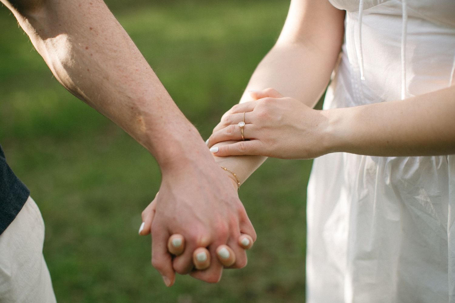 Close-up of a couple holding hands, woman wearing an engagement ring, with a green garden background.