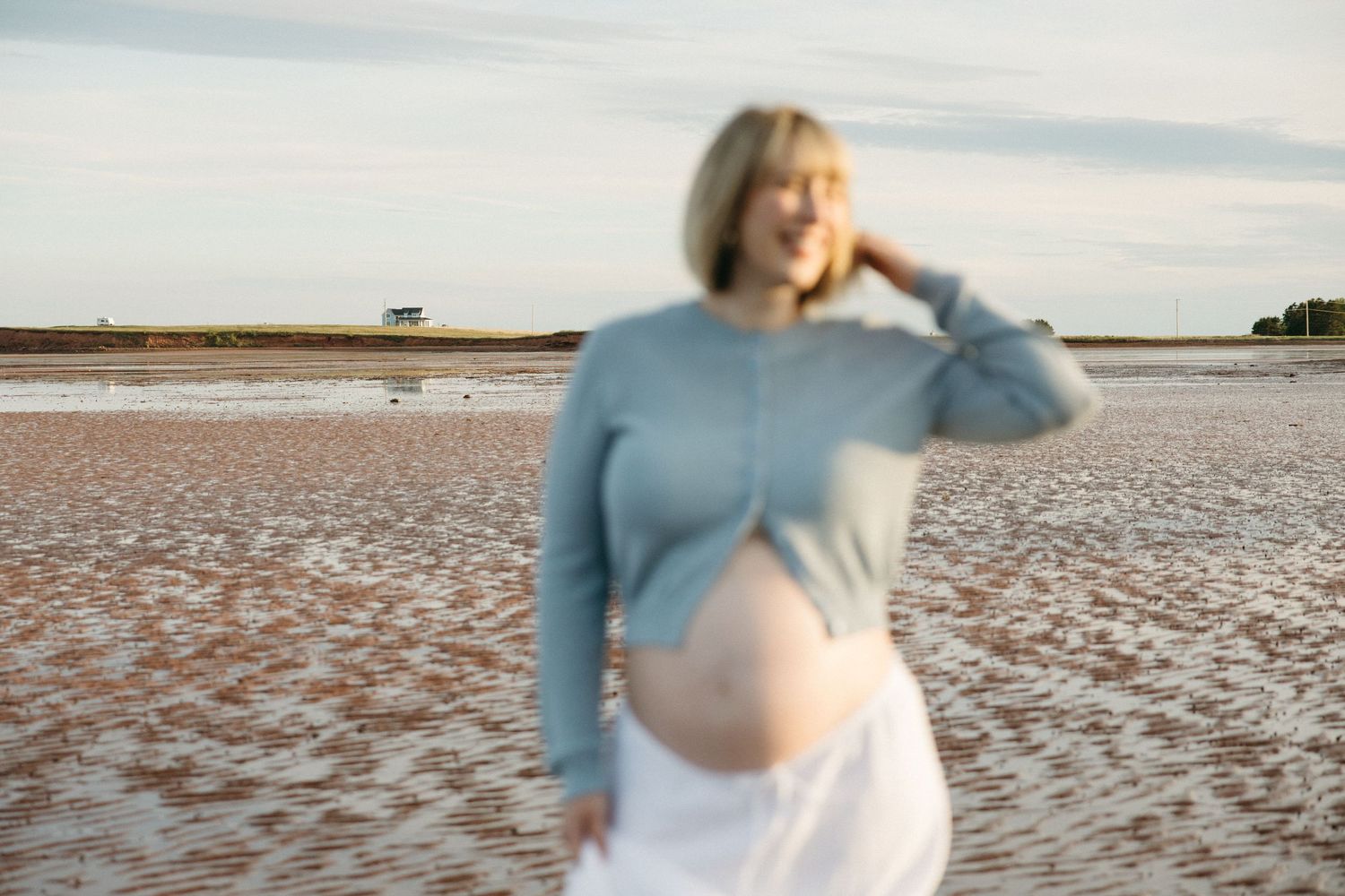 Smiling pregnant woman in blue sweater and white skirt standing on a sandy beach, cradling her baby bump.