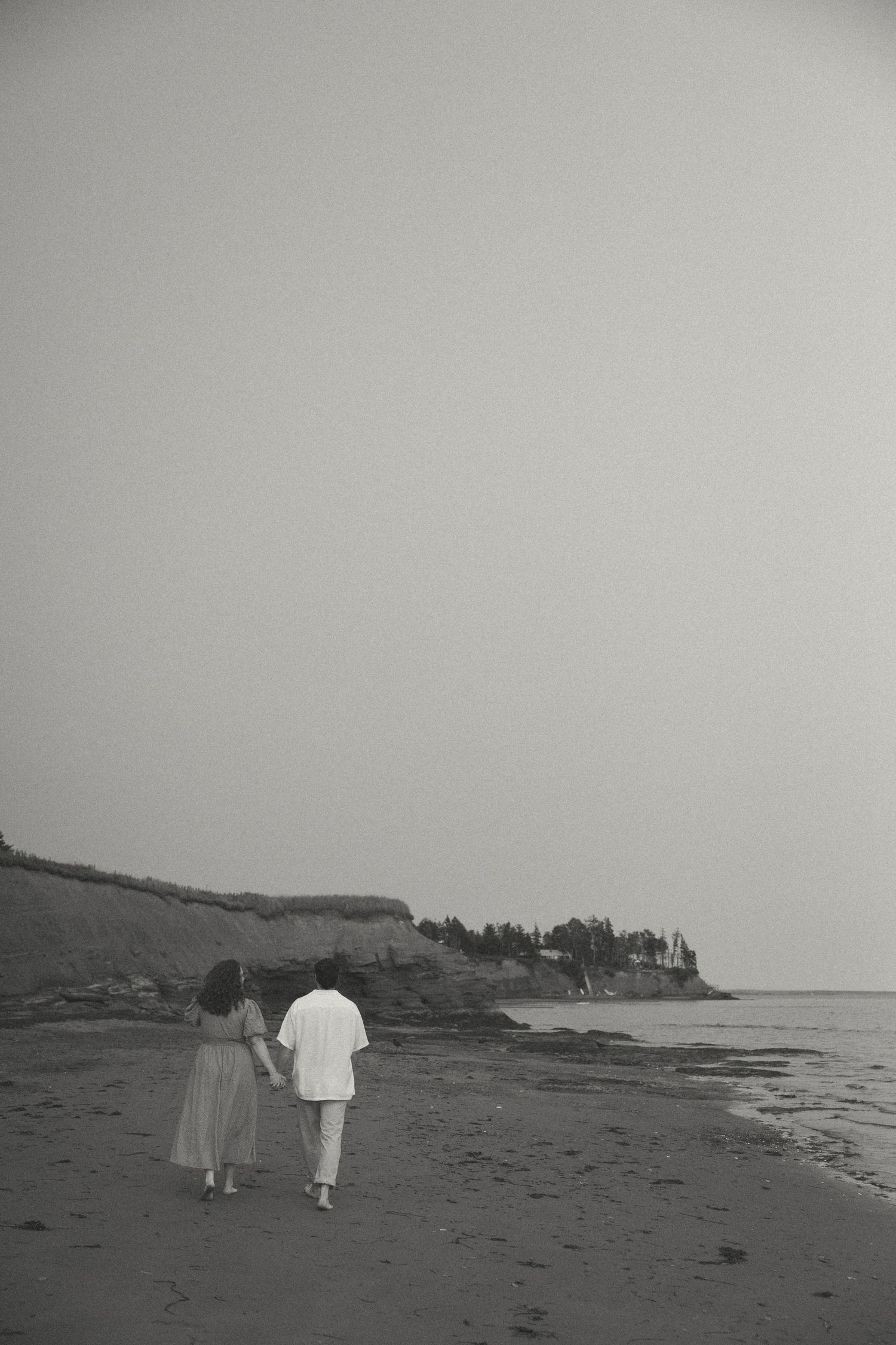 Black and white image of two figures walking along a serene beach shoreline.