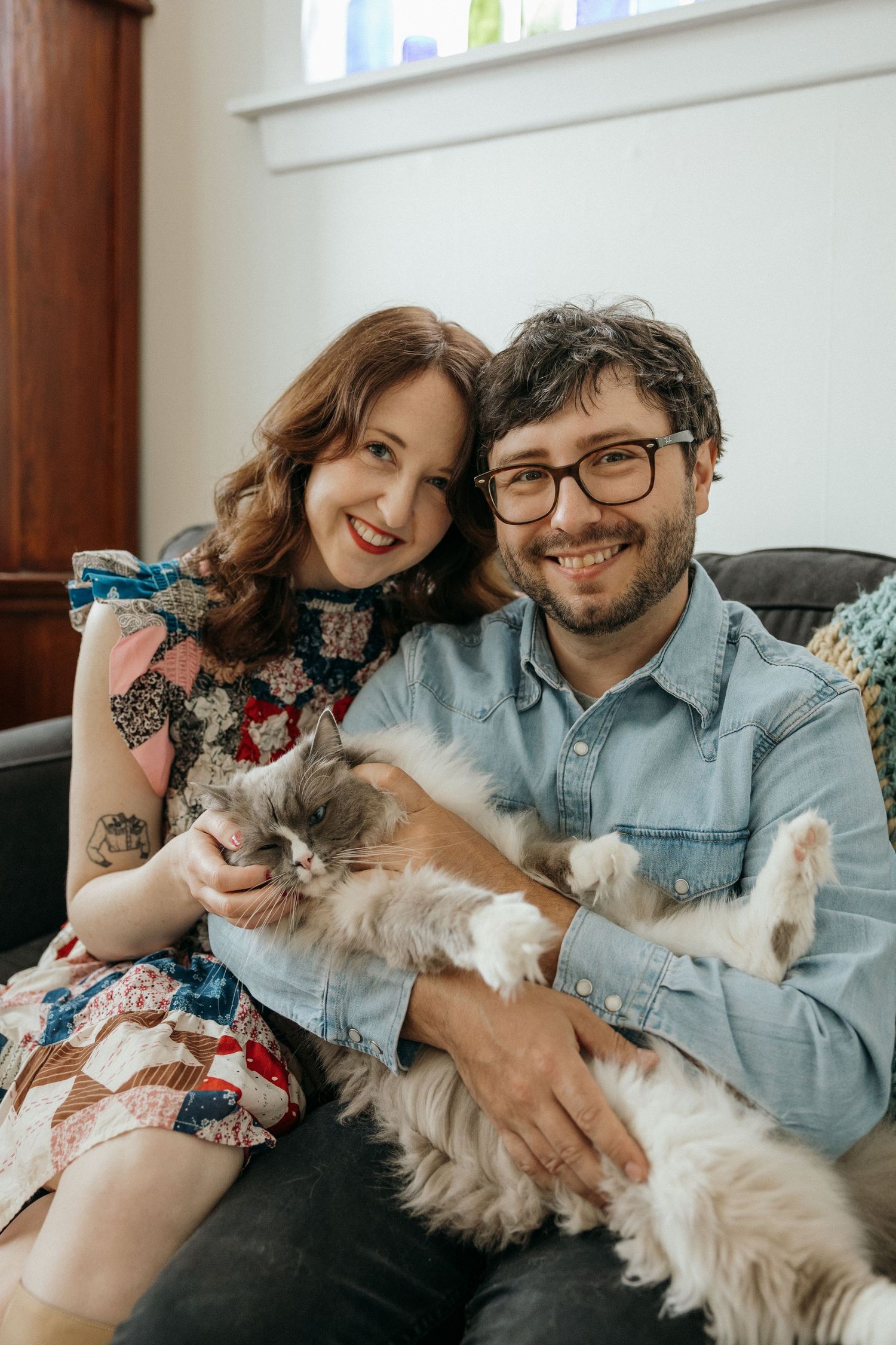 Couple snuggles with fluffy cats while reading on comfortable couch.