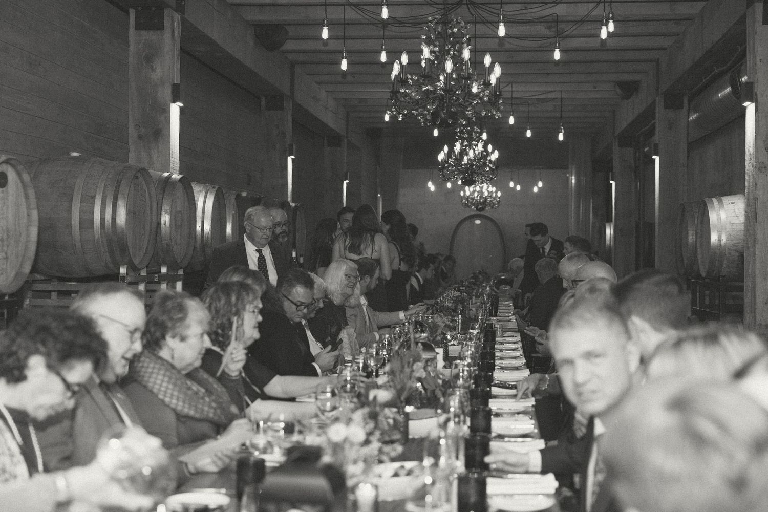 Wedding reception guests enjoying dinner and drinks in a rustic venue with wooden beams and string lights.