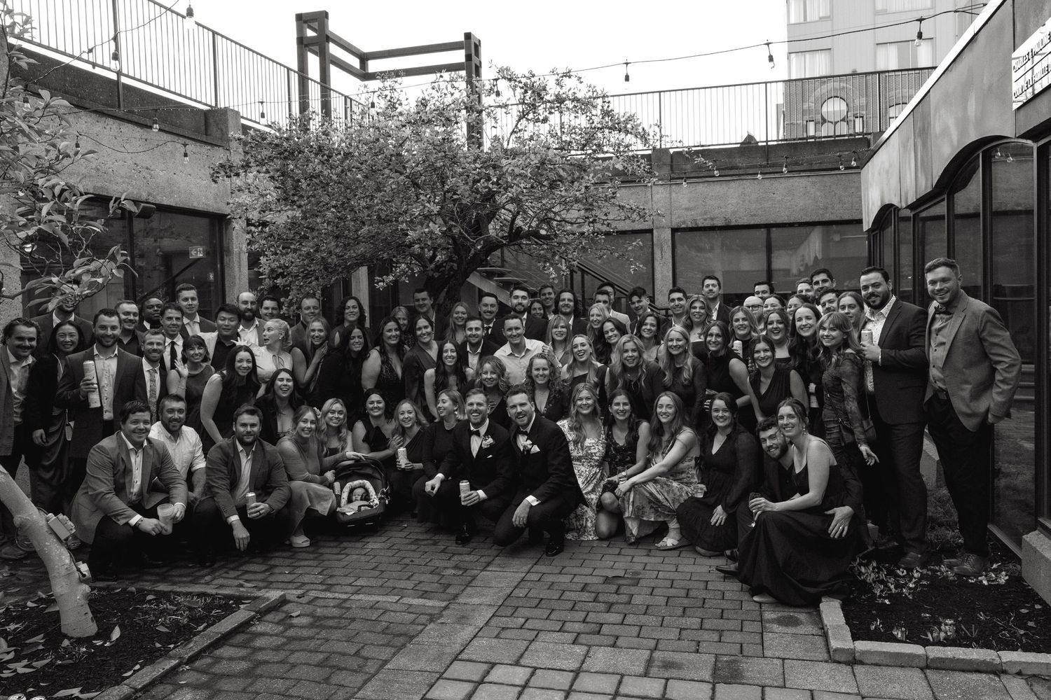 Large group photo of wedding guests gathered in an outdoor courtyard with brick pavement.