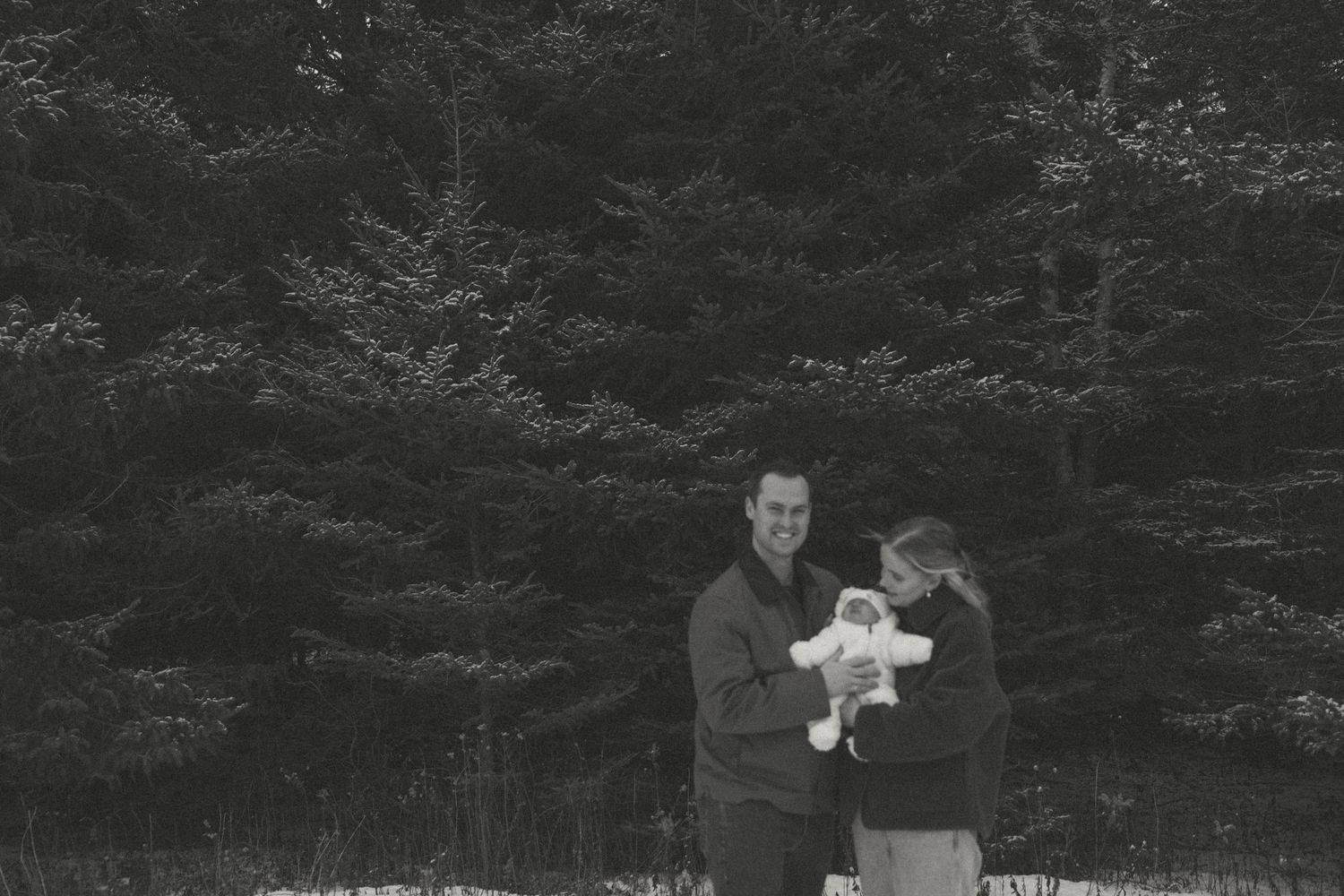 A black and white photo of a couple with their baby against a backdrop of evergreen trees.