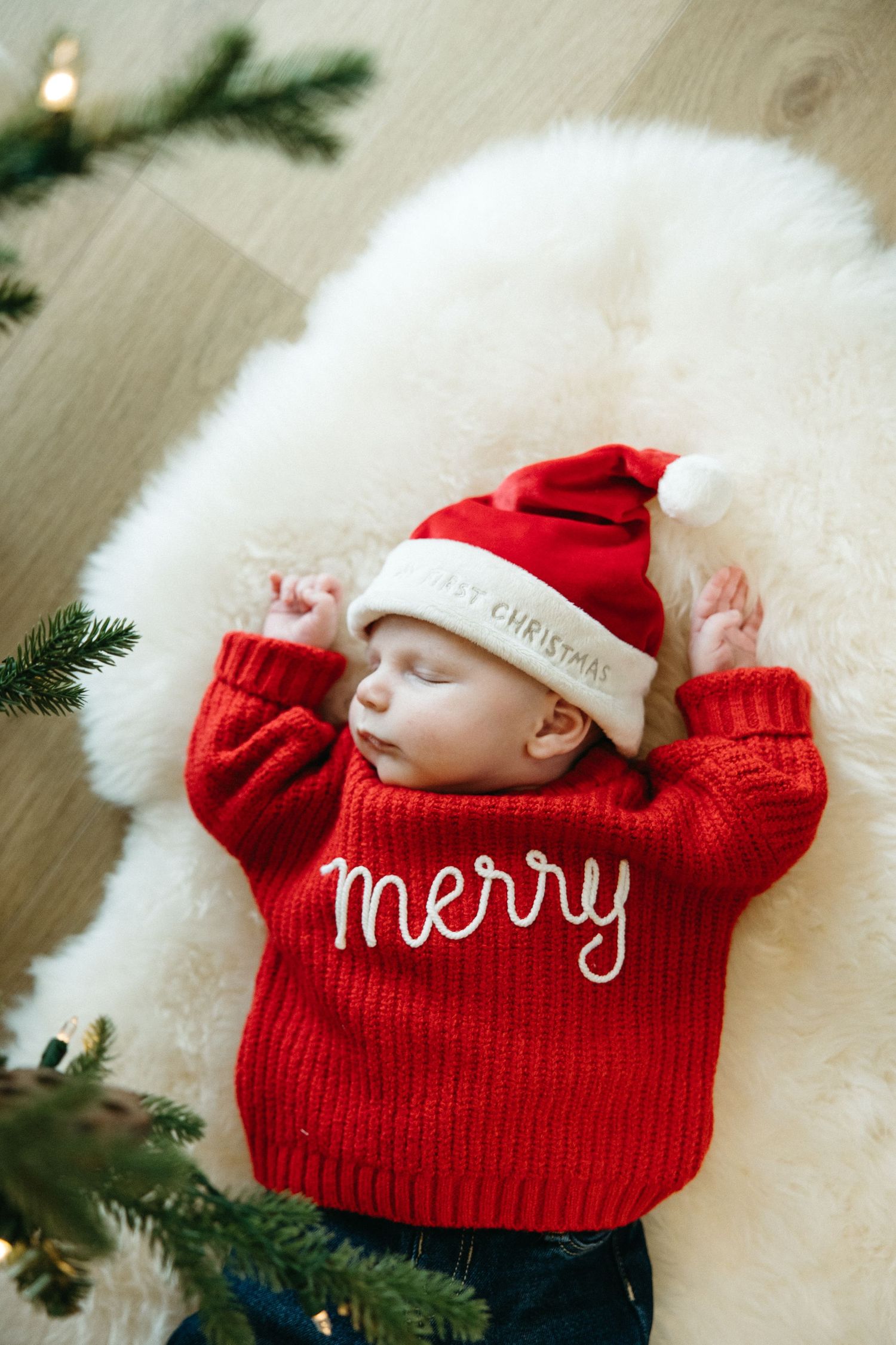 A peaceful baby wearing a red Christmas sweater and Santa hat rests on a white fur rug.