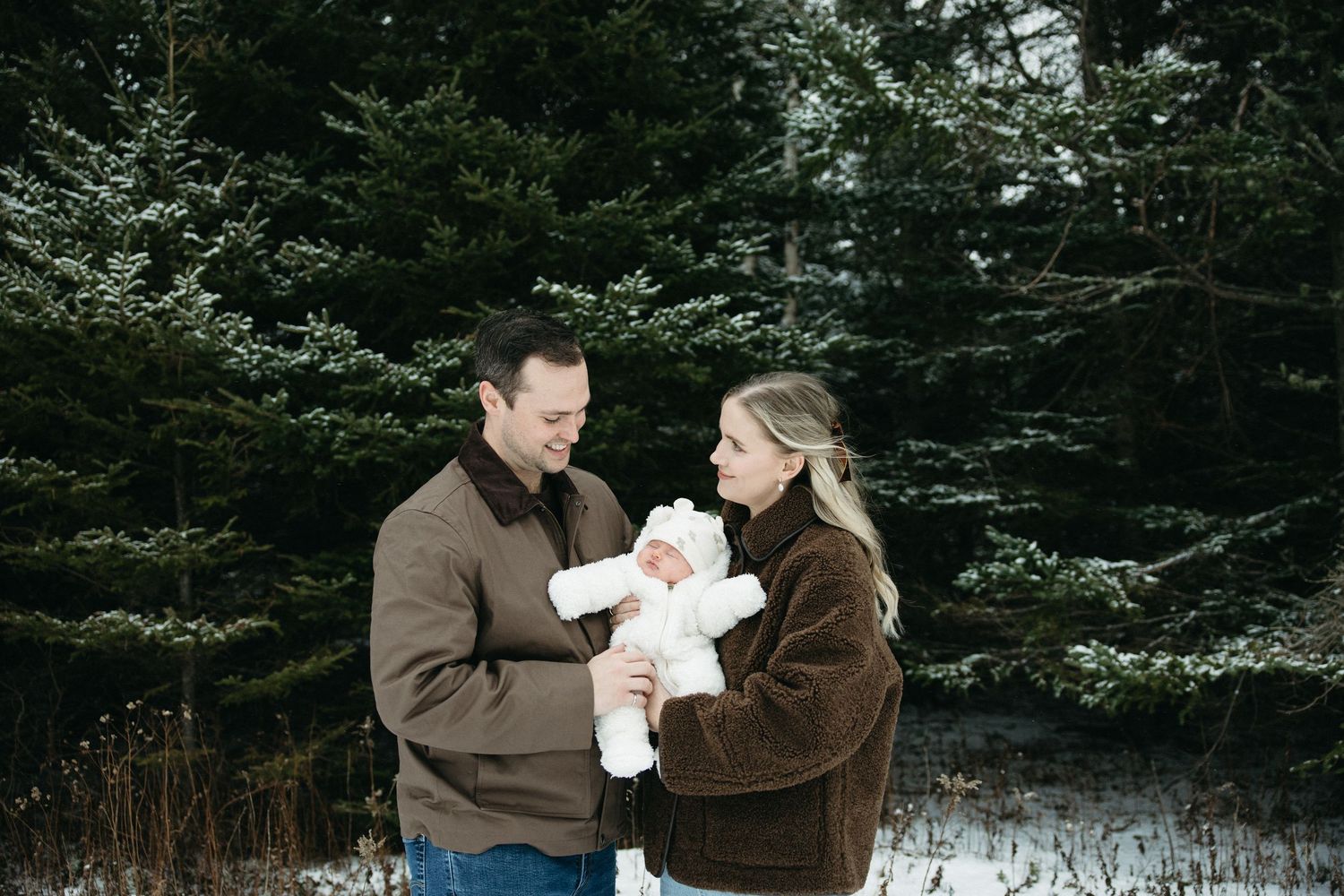 A family embraces in a snowy winter setting surrounded by evergreen trees.