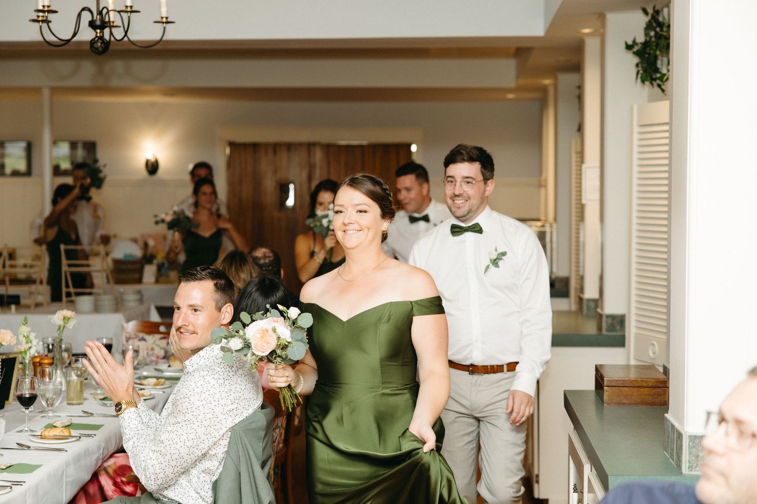 Bridesmaid in green dress enters wedding reception hall carrying white bouquet with groom nearby.