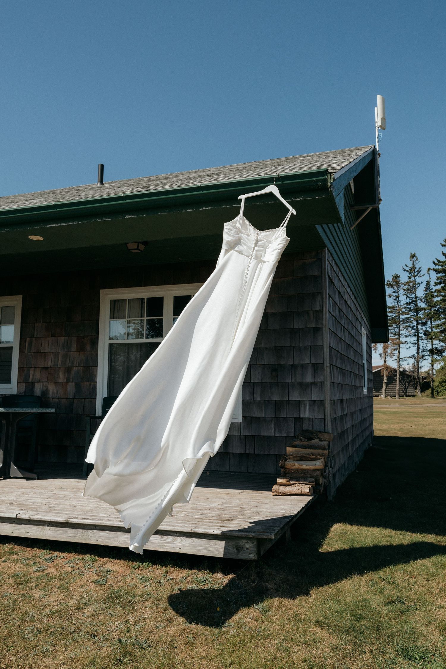 White wedding dress hanging on a hanger outside a rustic gray shingle cottage on a sunny day.