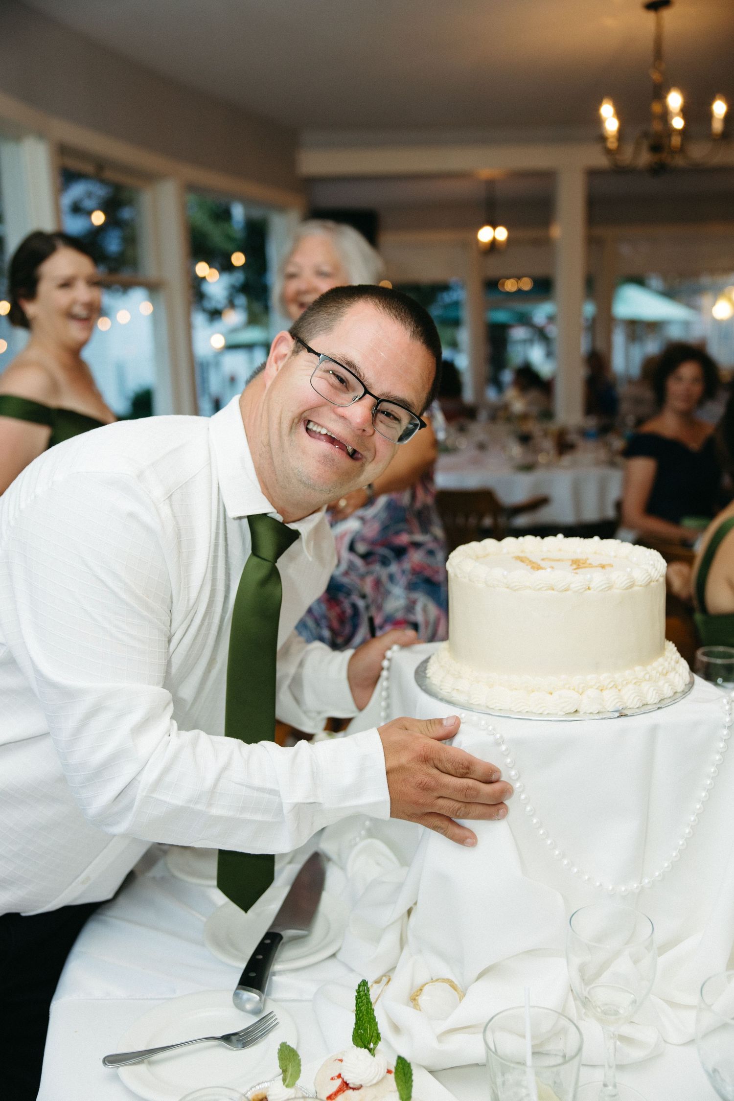 Smiling groom with glasses and green tie hugging wedding cake at reception.