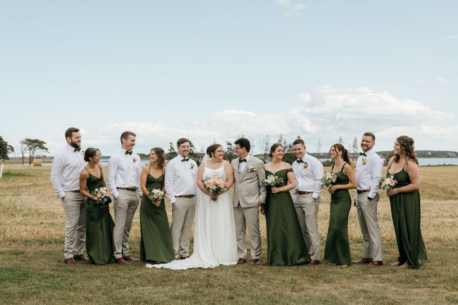 Wedding party in green bridesmaid dresses and tan suits posing in an open field by the water.