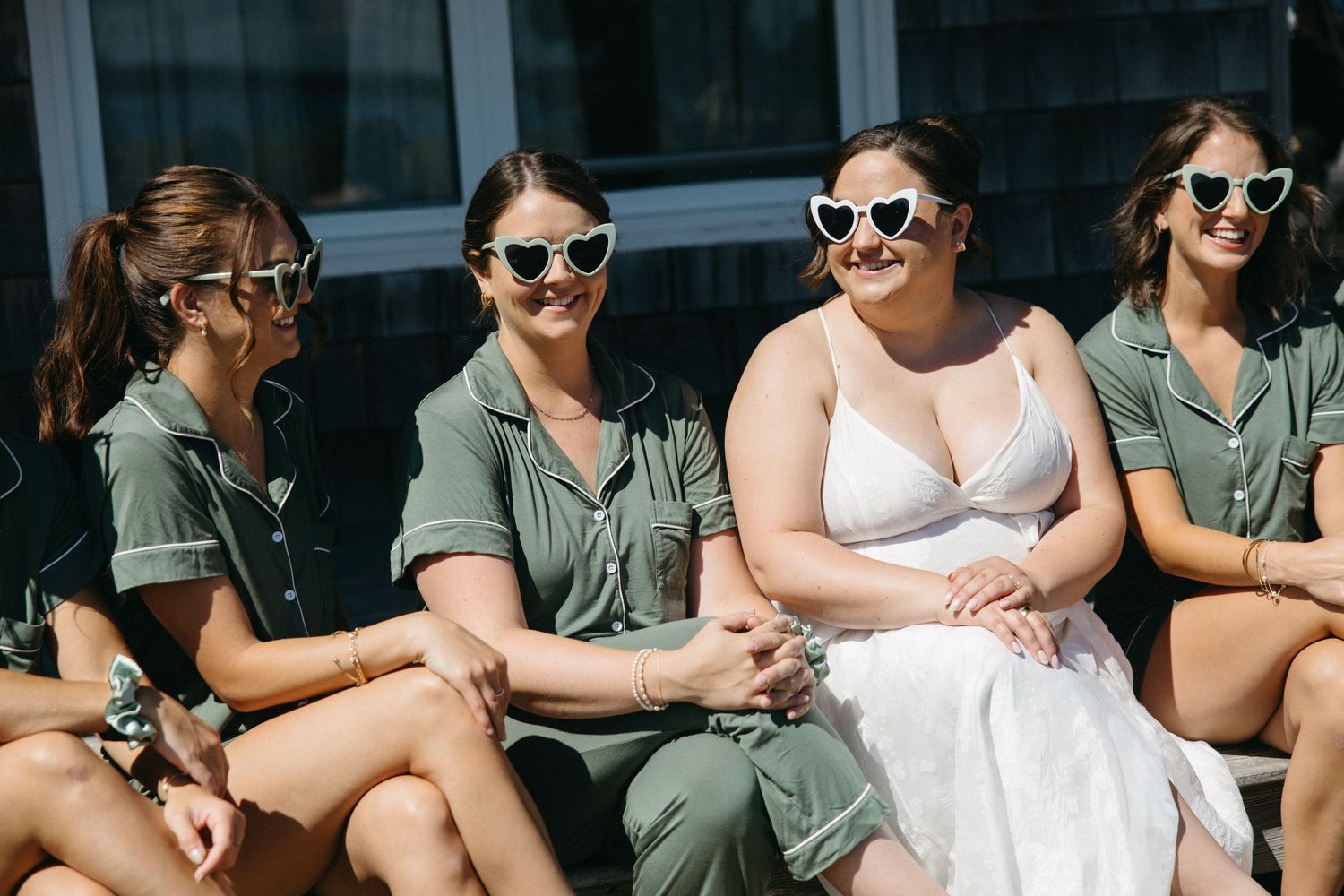 Bride laughing with bridesmaids in matching olive green outfits and heart-shaped sunglasses on a sunny wedding day.