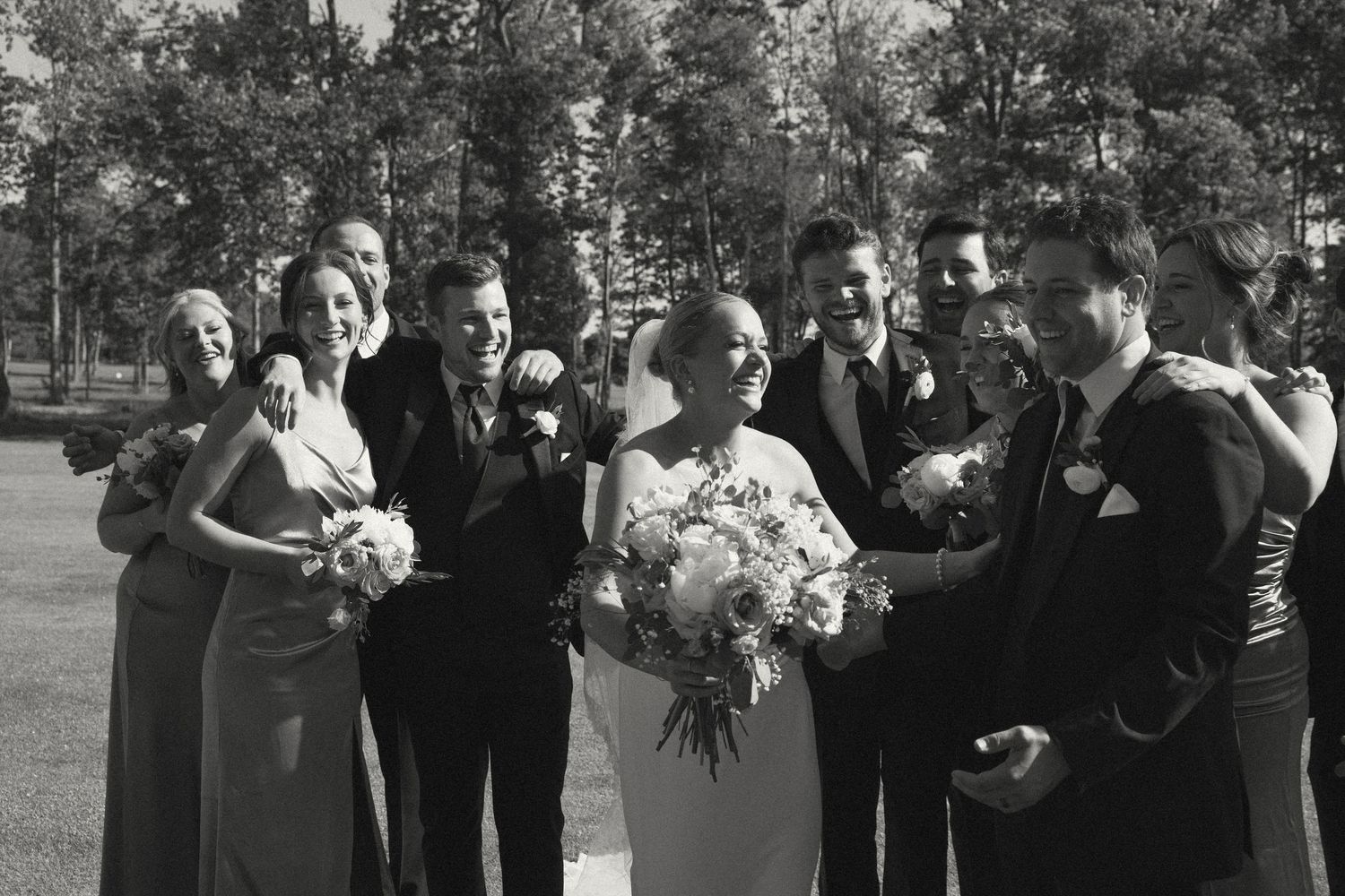 Wedding party gathers outdoors with bouquets against a backdrop of pine trees.