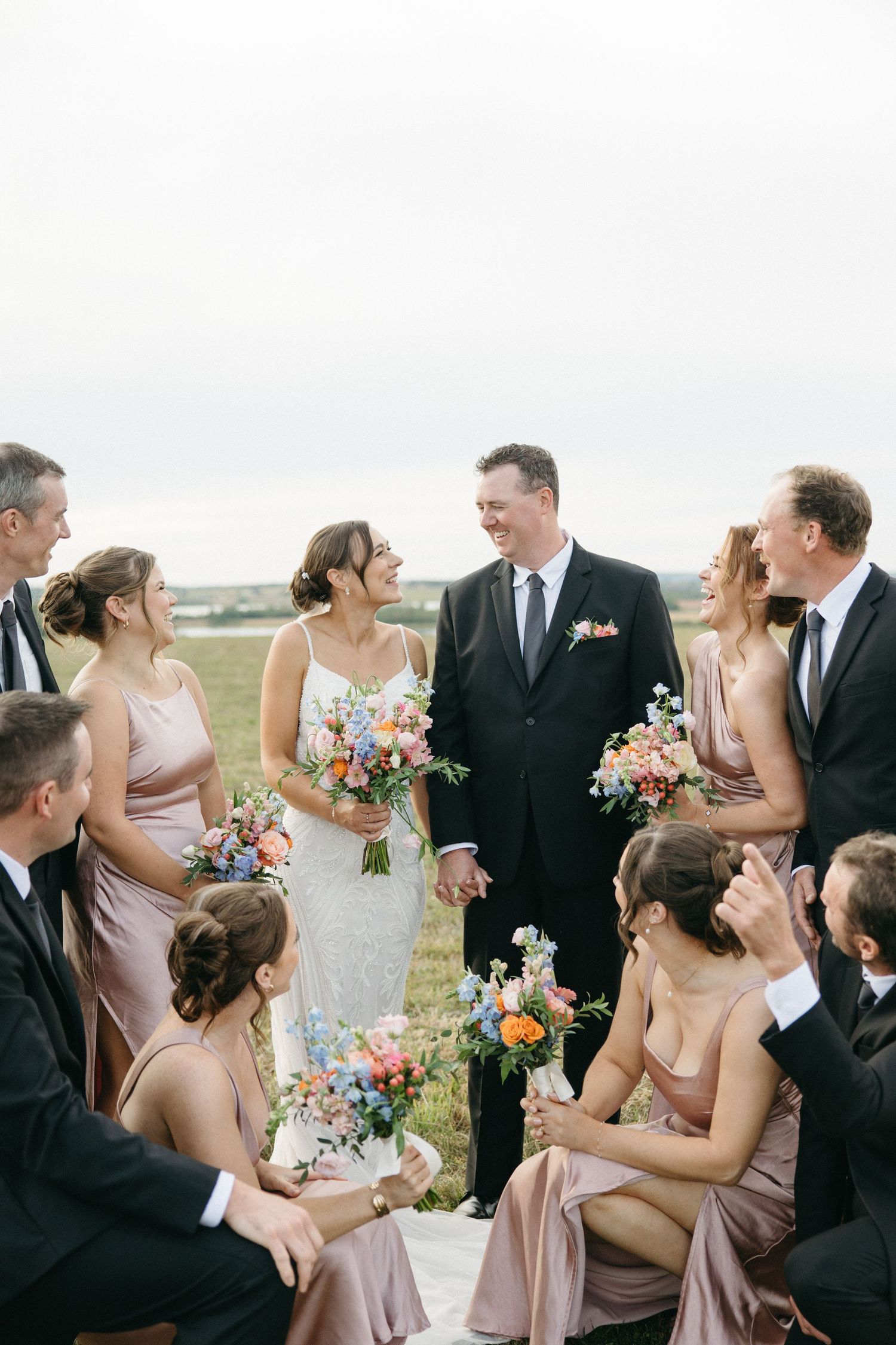 Wedding party members interact and laugh together while holding colorful bouquets outdoors.