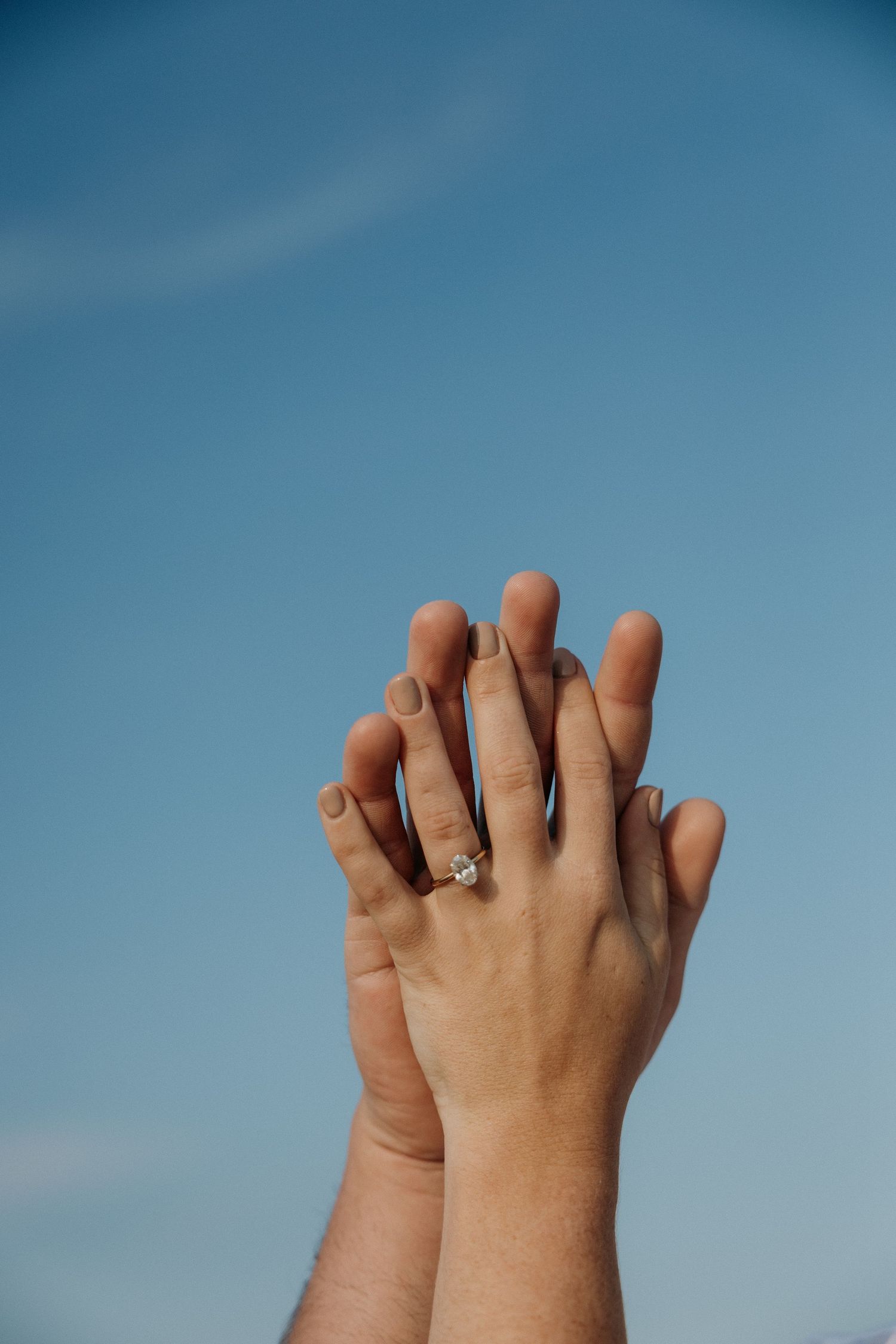 Two hands intertwined against a blue sky, showcasing an engagement ring.