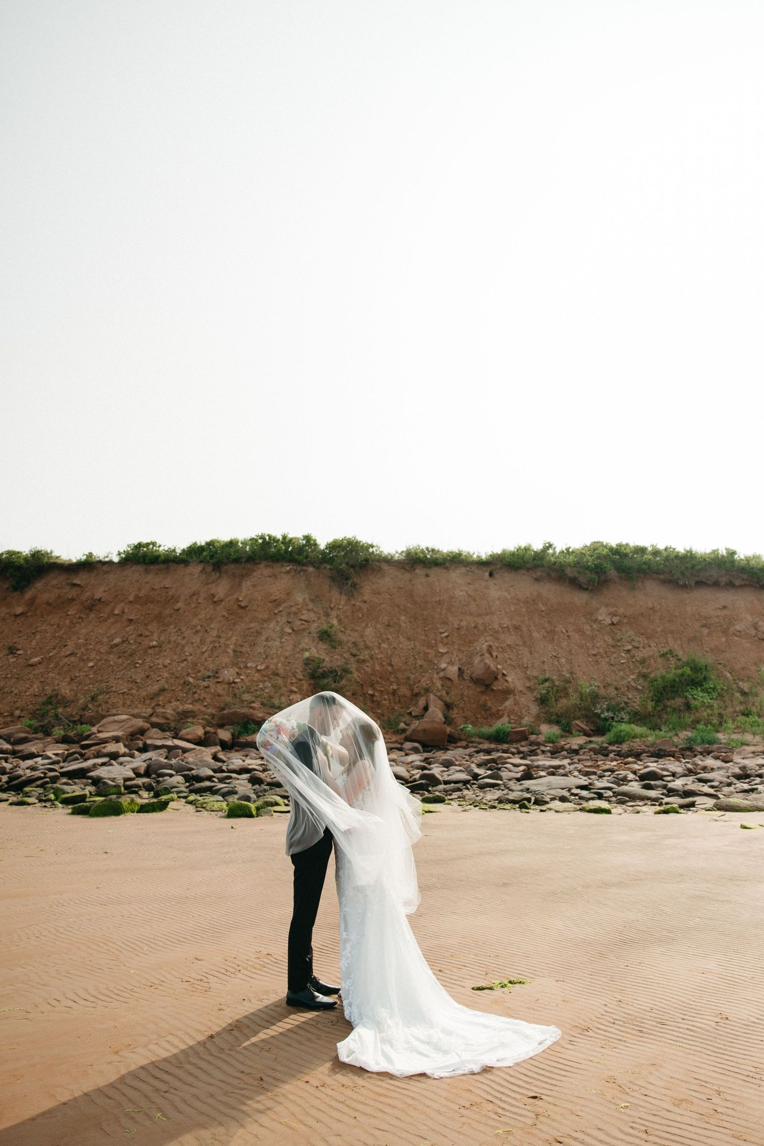 Wedding couple photographed from behind on sandy beach with dramatic cliffside backdrop.